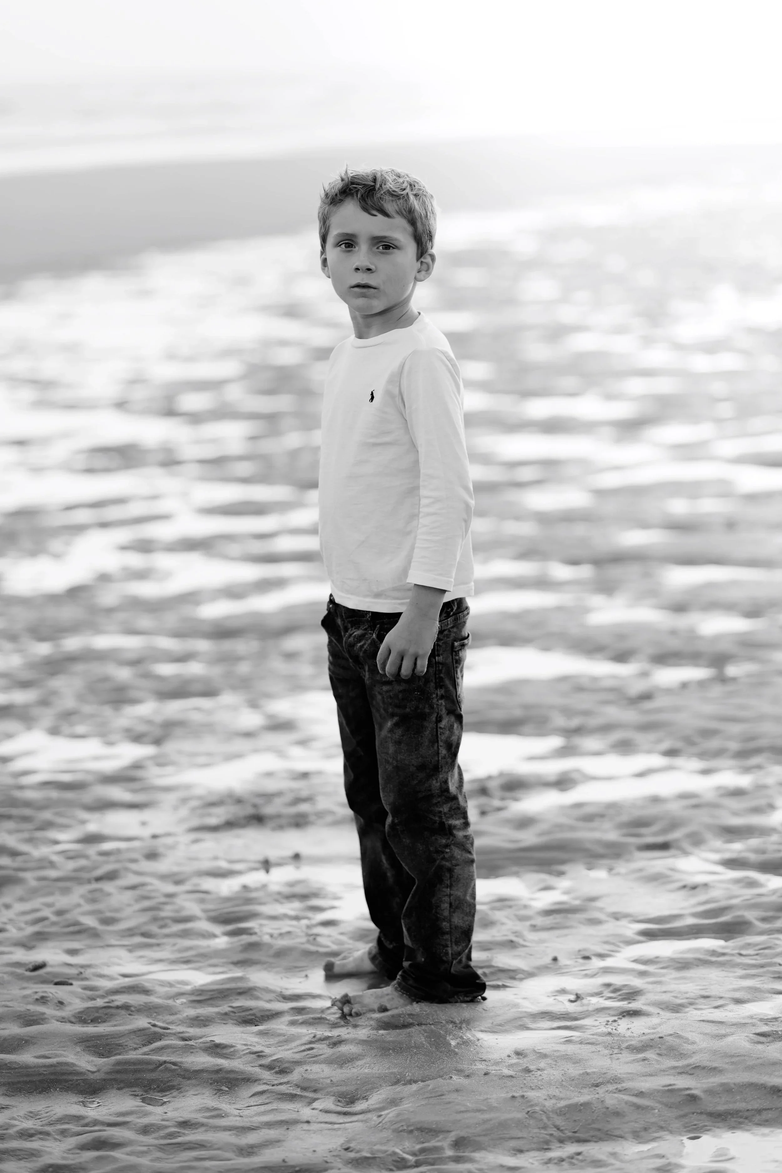 A young boy standing on a sandy beach with water in the background, looking at the camera.