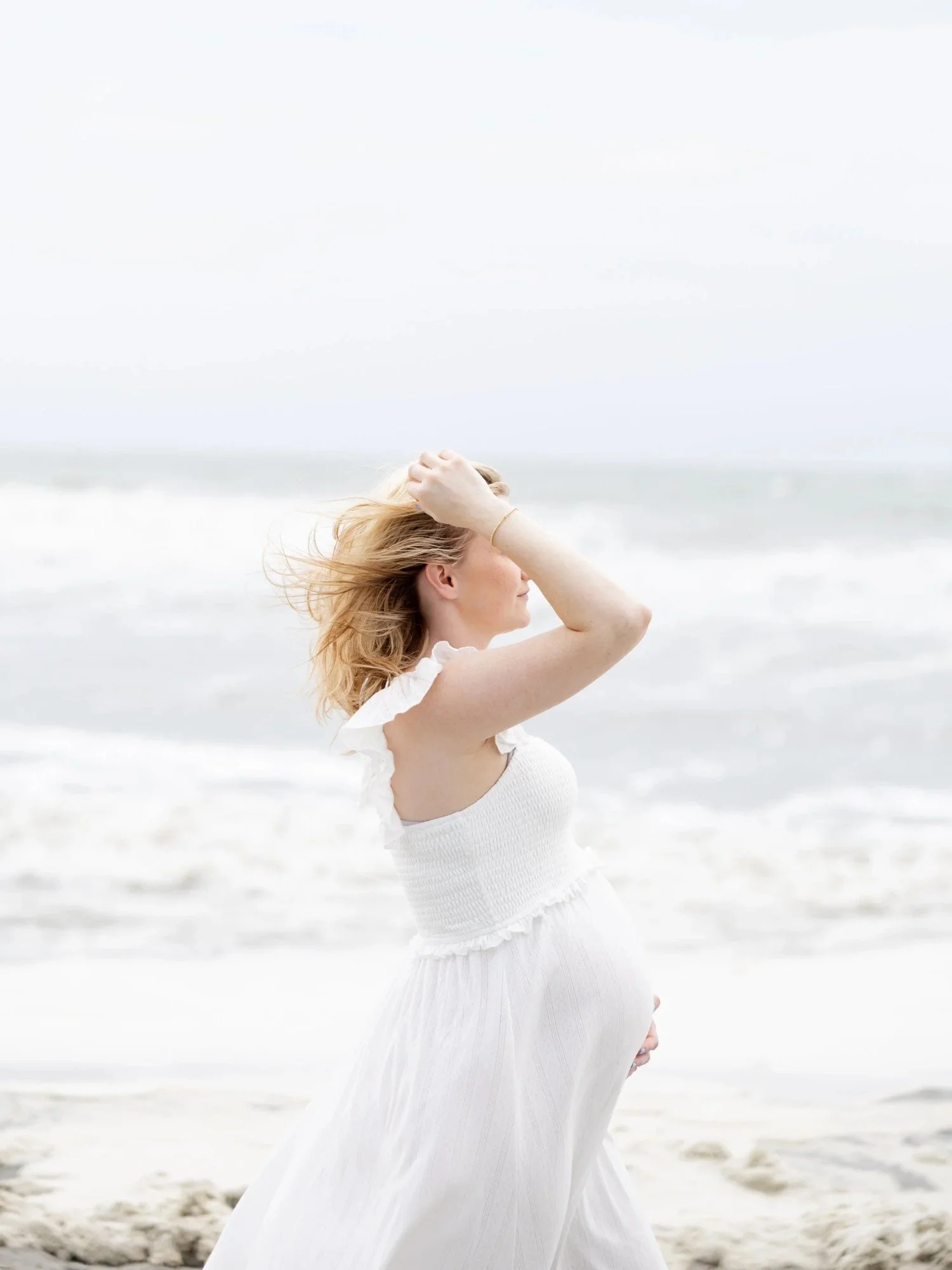 A pregnant woman in a white dress standing on the beach, holding her hair with one hand and resting her other hand on her belly, with the ocean in the background.