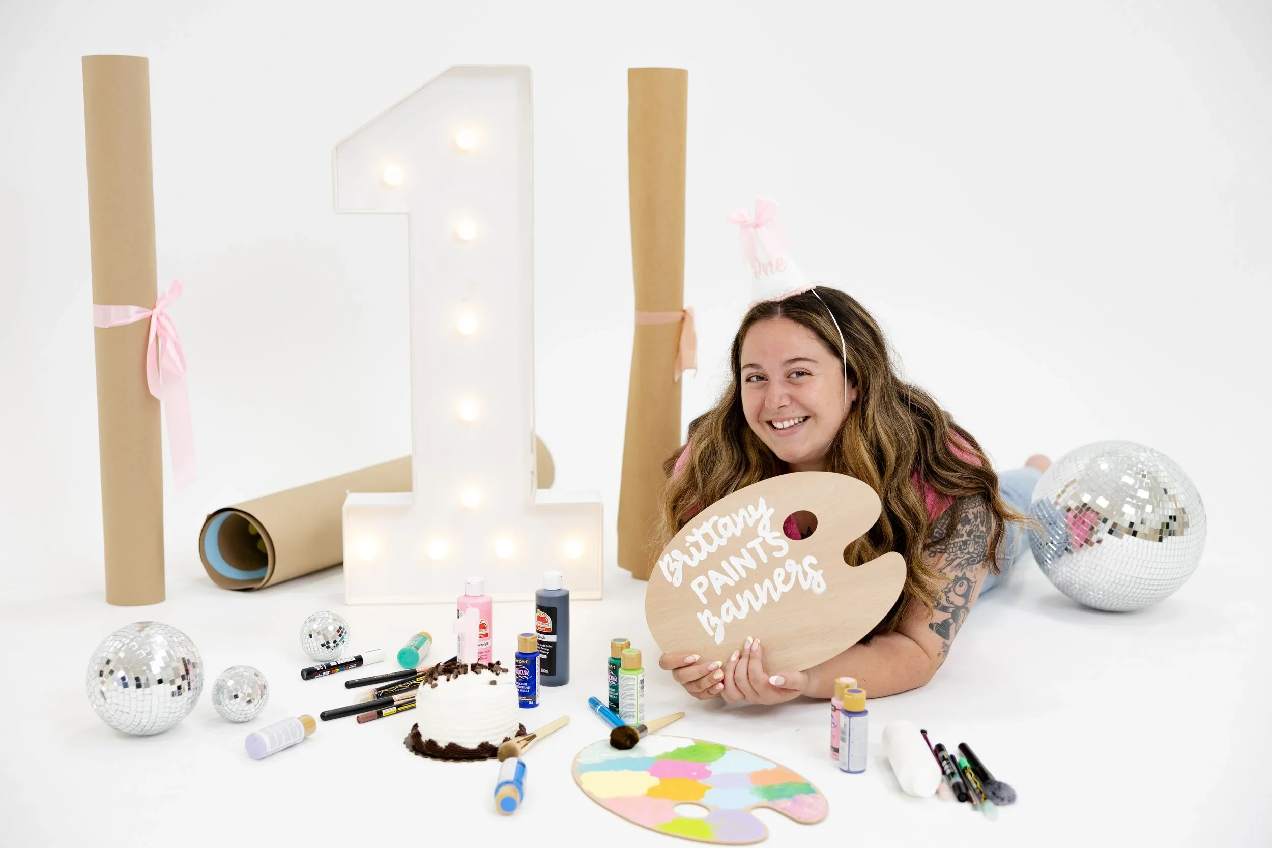 A girl with long hair and tattoos lying on her stomach surrounded by art supplies, disco balls, and party decorations with a large illuminated number one in the background.