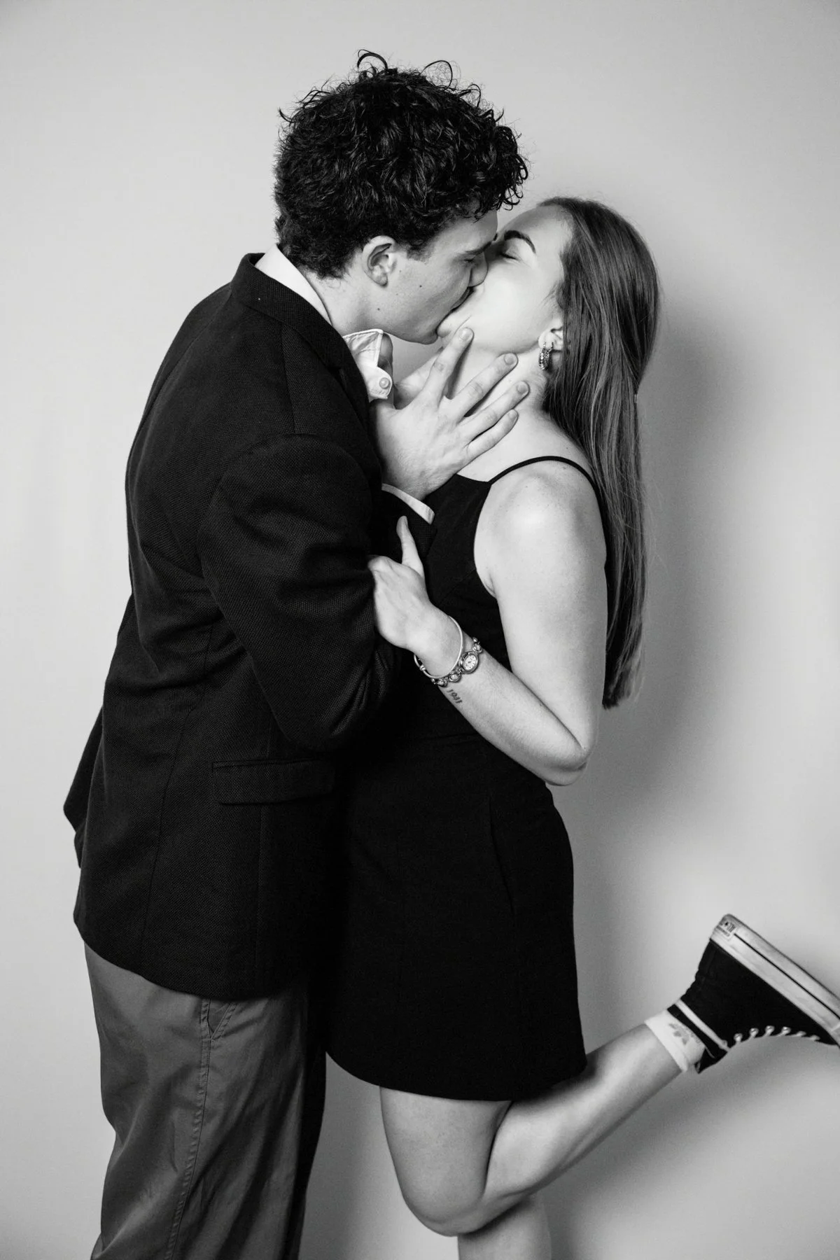A black and white photo of a young man and woman kissing. The man, dressed in a suit jacket and shirt, is holding the woman's face while she, in a sleeveless dress, is on one leg with her foot up. She is wearing sneakers, and accessories including ea