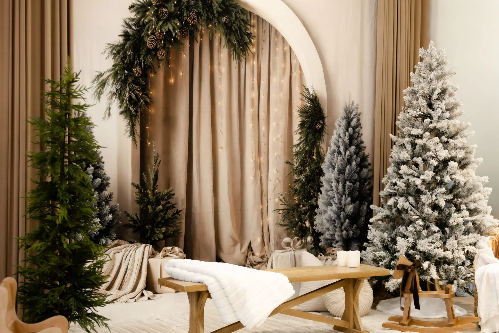 Christmas decorated room with white snowy Christmas trees, garlands made of pine branches and pinecones, and warm lighting. A cozy seating area with blankets, candles, and a wooden table.