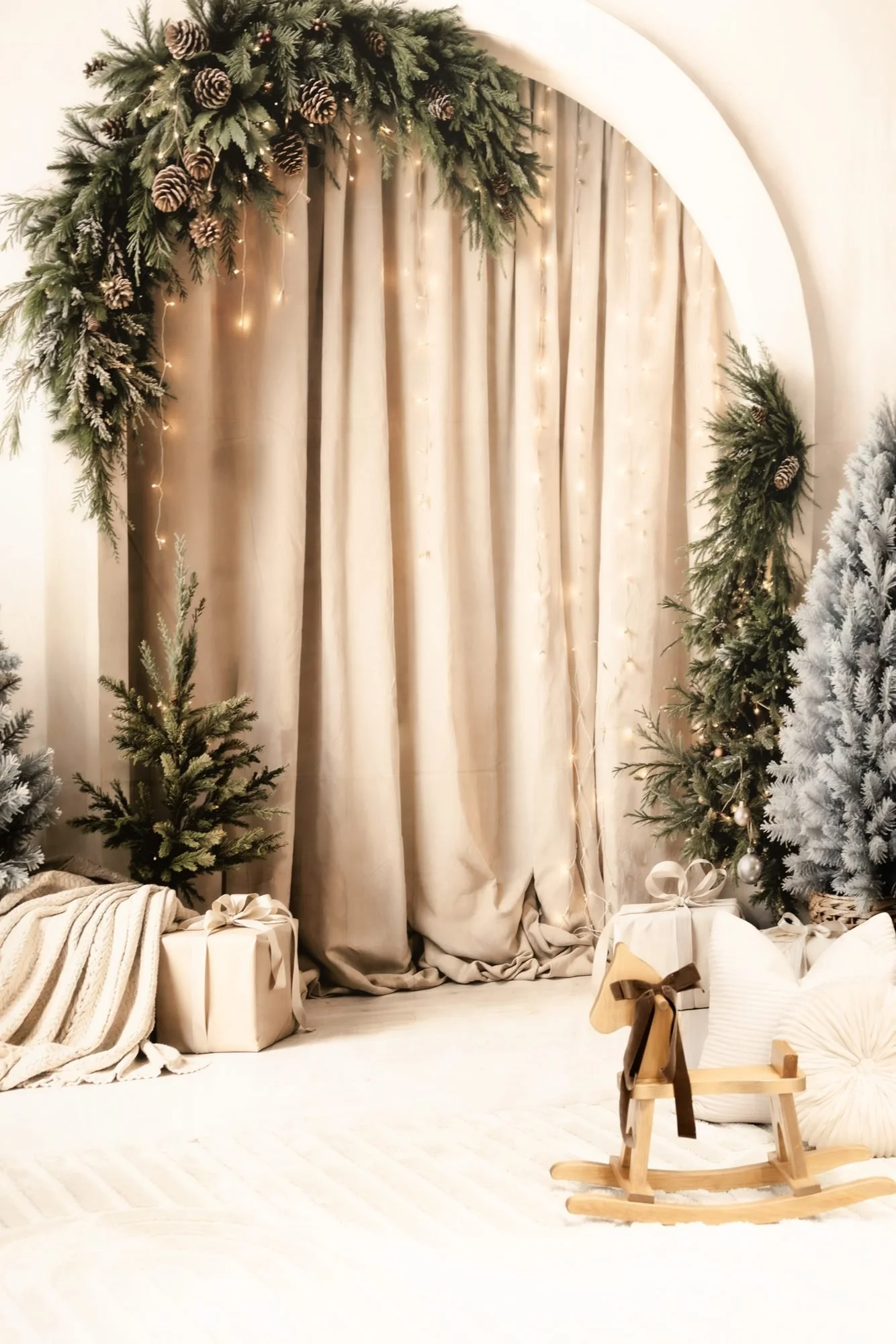 Christmas-themed scene with a beige curtain backdrop, decorated with pine branches and pinecones. Wrapped gifts and a blanket are on the floor, with a small wooden rocking horse and a white pillow creating a cozy setting.
