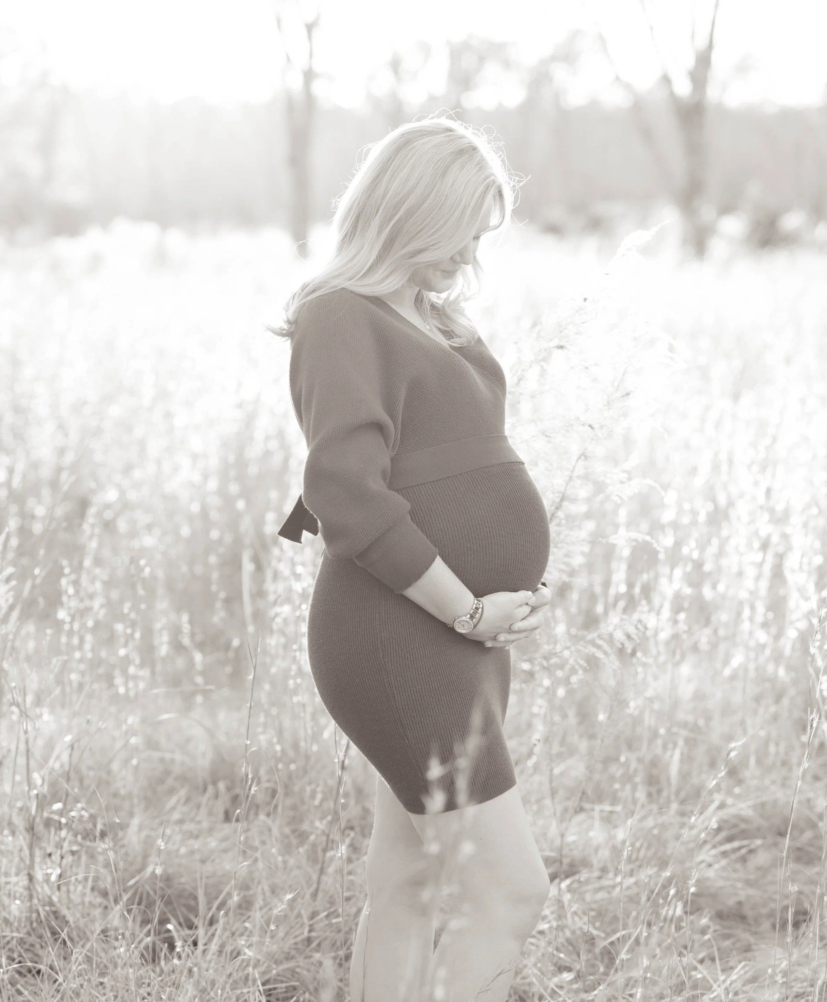A woman with long blonde hair cradling her pregnant belly in a rural outdoor setting with tall dry grass and trees in the background, captured in a black-and-white photograph.