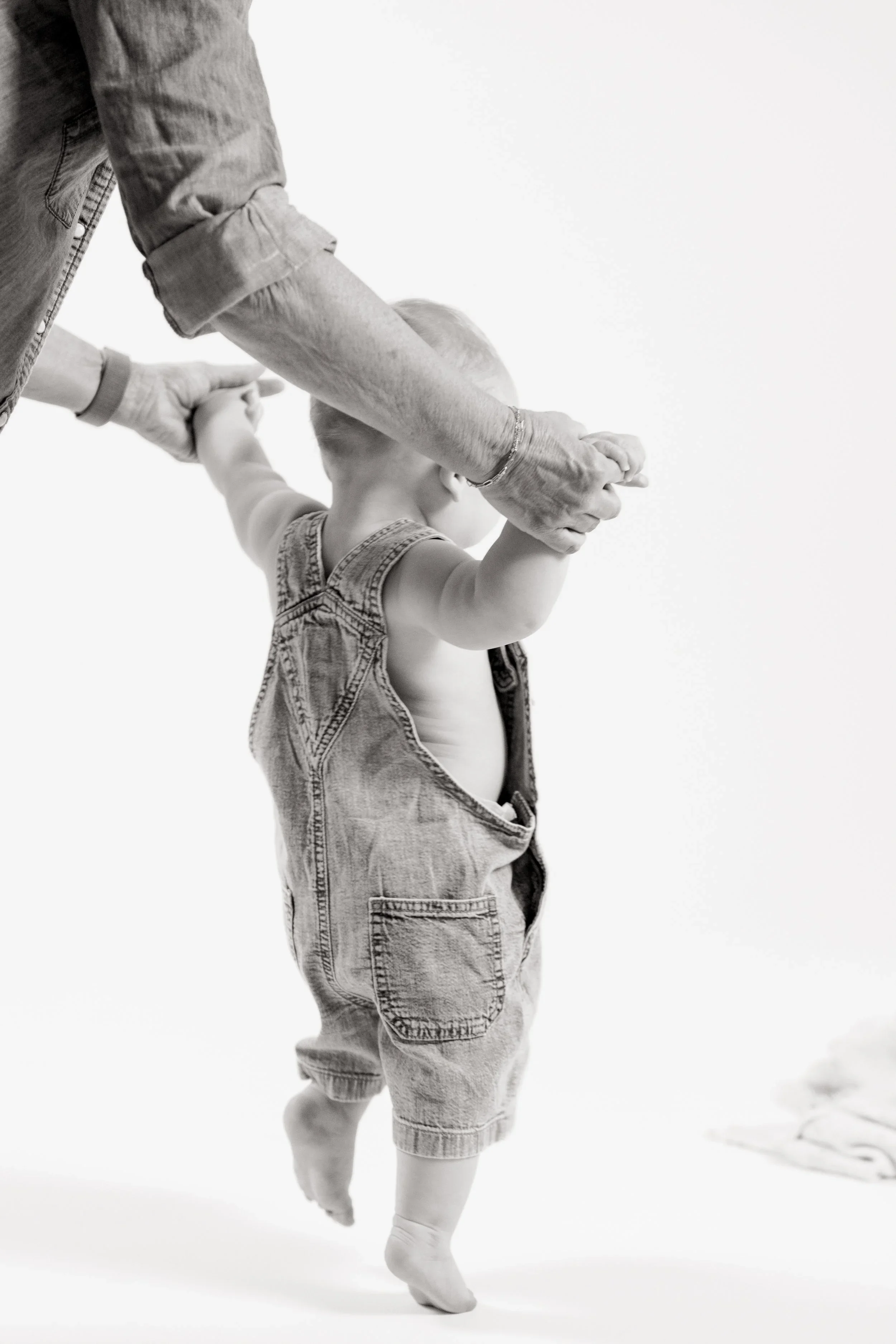 A child in denim overalls walking with assistance from an adult, holding hands, against a plain white background.