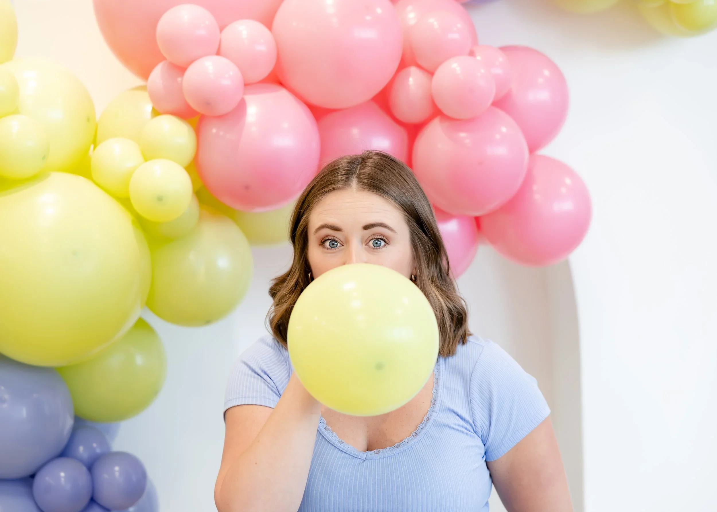 A woman with blue eyes and brown hair holding a yellow balloon in front of her face, surrounded by pink, yellow, purple, and blue balloons.
