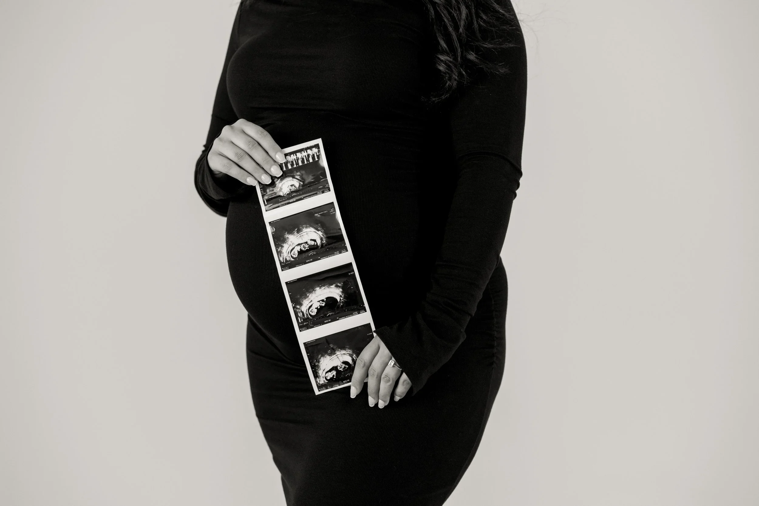 A pregnant woman in a black dress holding ultrasound images in front of her belly.