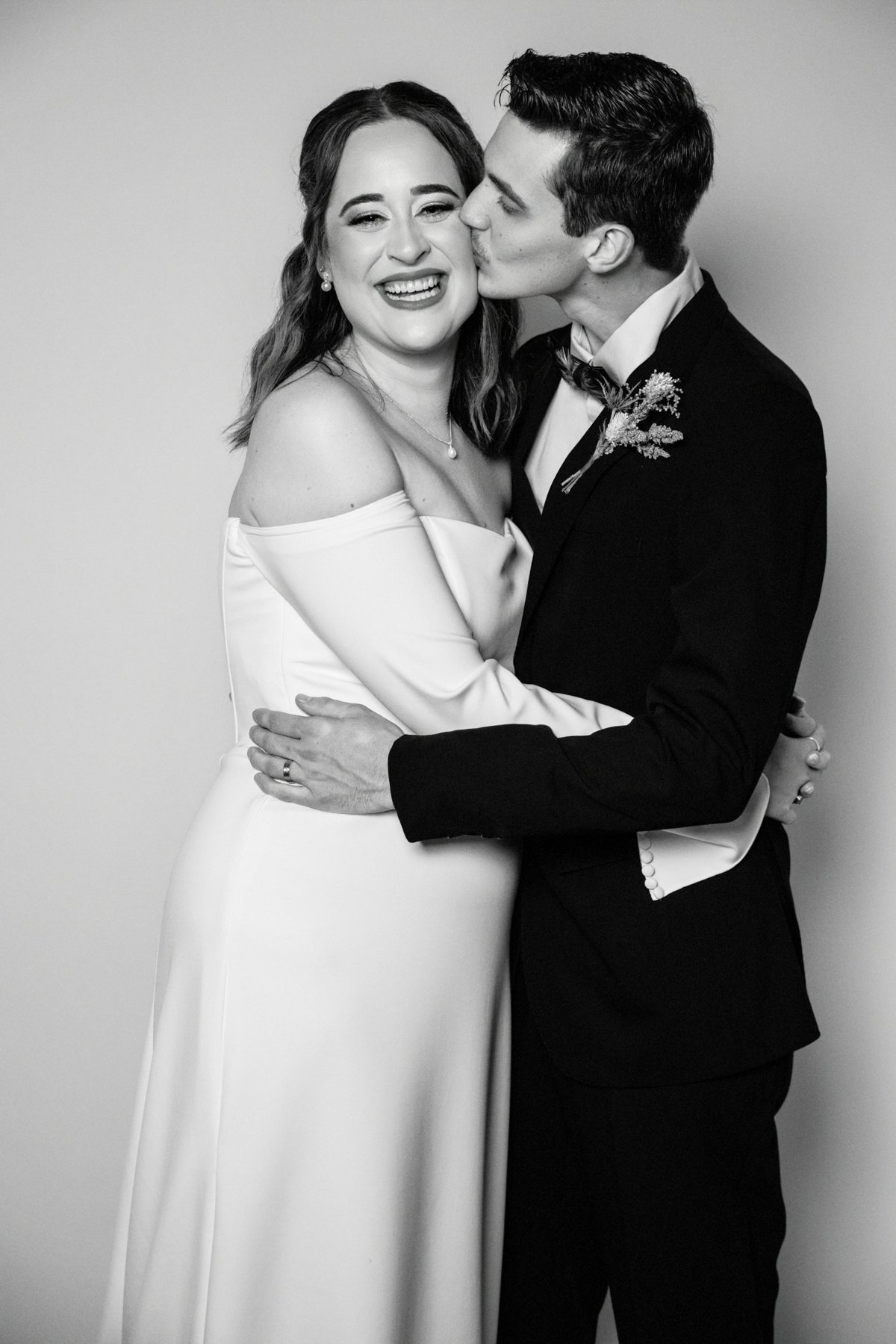Black and white photo of a couple in wedding attire, embracing and smiling, with the man giving a kiss on the woman's cheek.