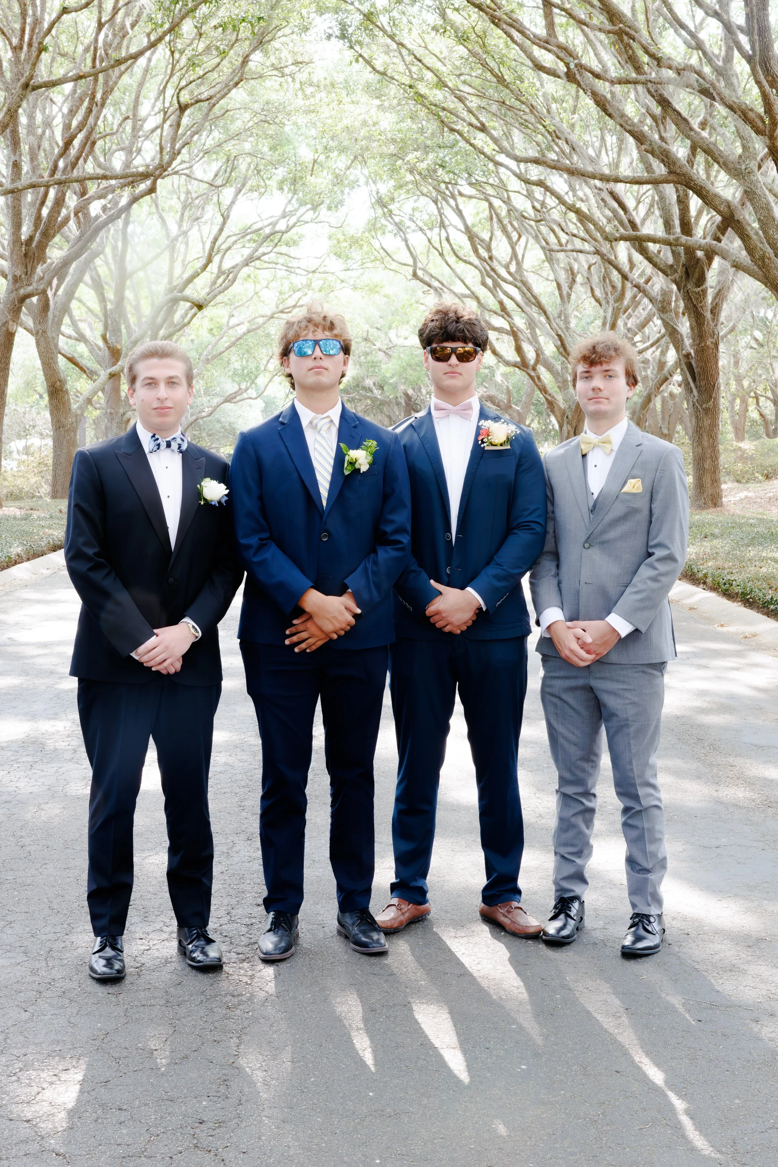 Four young men in suits standing outdoors on a paved path under trees, facing forward, dressed in formal attire with boutonnières and bow ties, some wearing sunglasses.