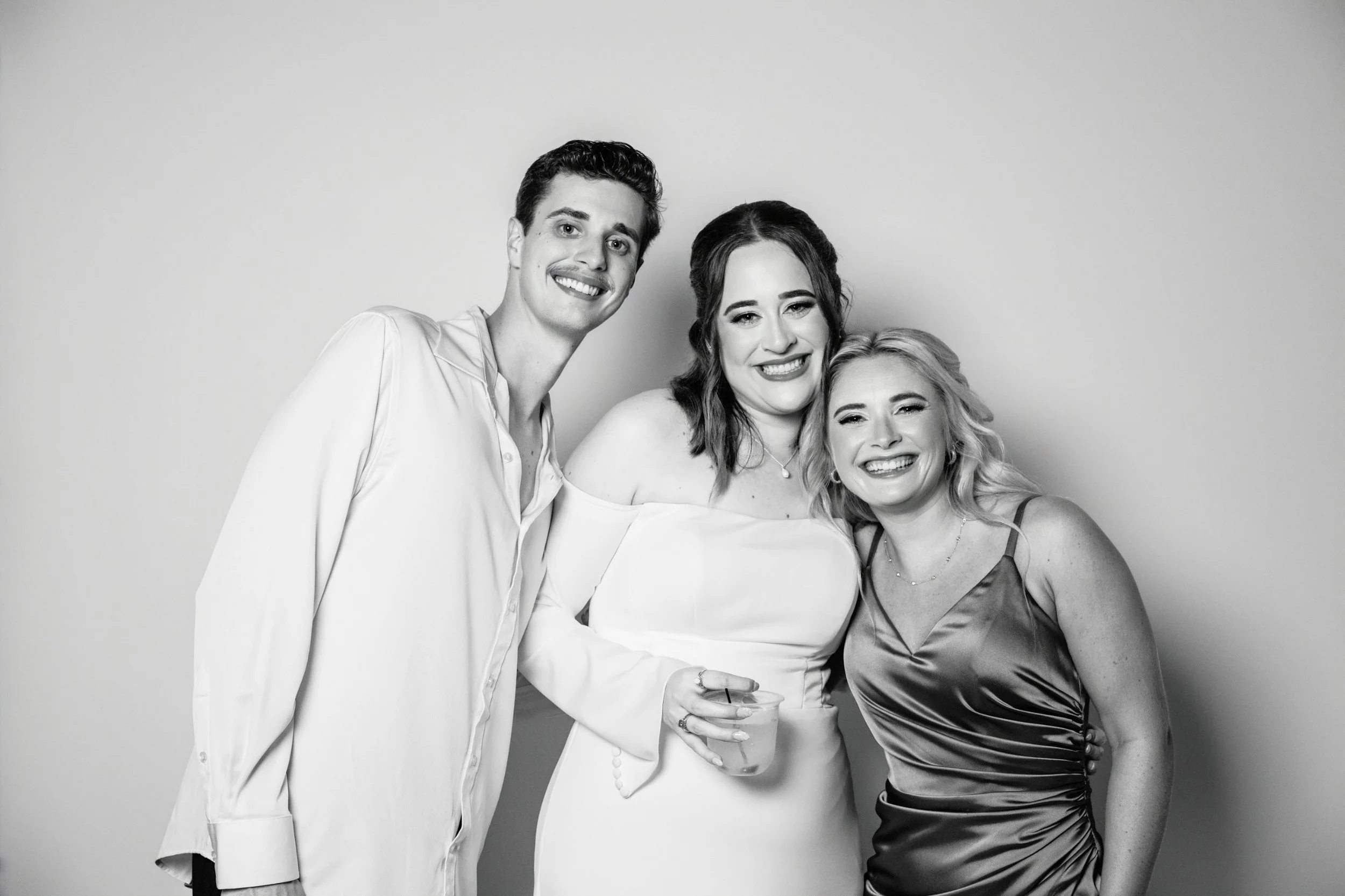Three people smiling and standing close together; one man in a white shirt, two women, one in a white dress and the other in a satin evening gown, holding a drink, against a plain wall.
