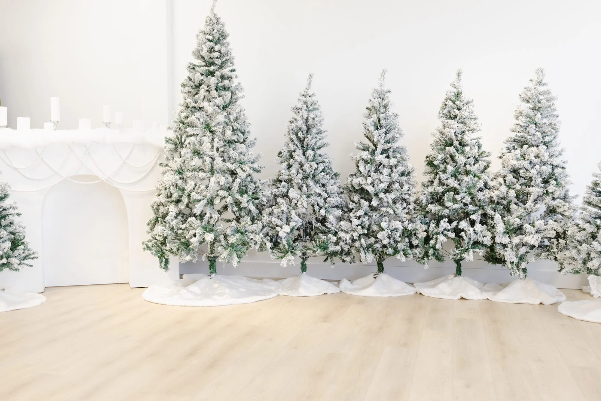 Decorative artificial snow-covered Christmas trees arranged in front of a white wall, with white drapes and candles on a table in the background.