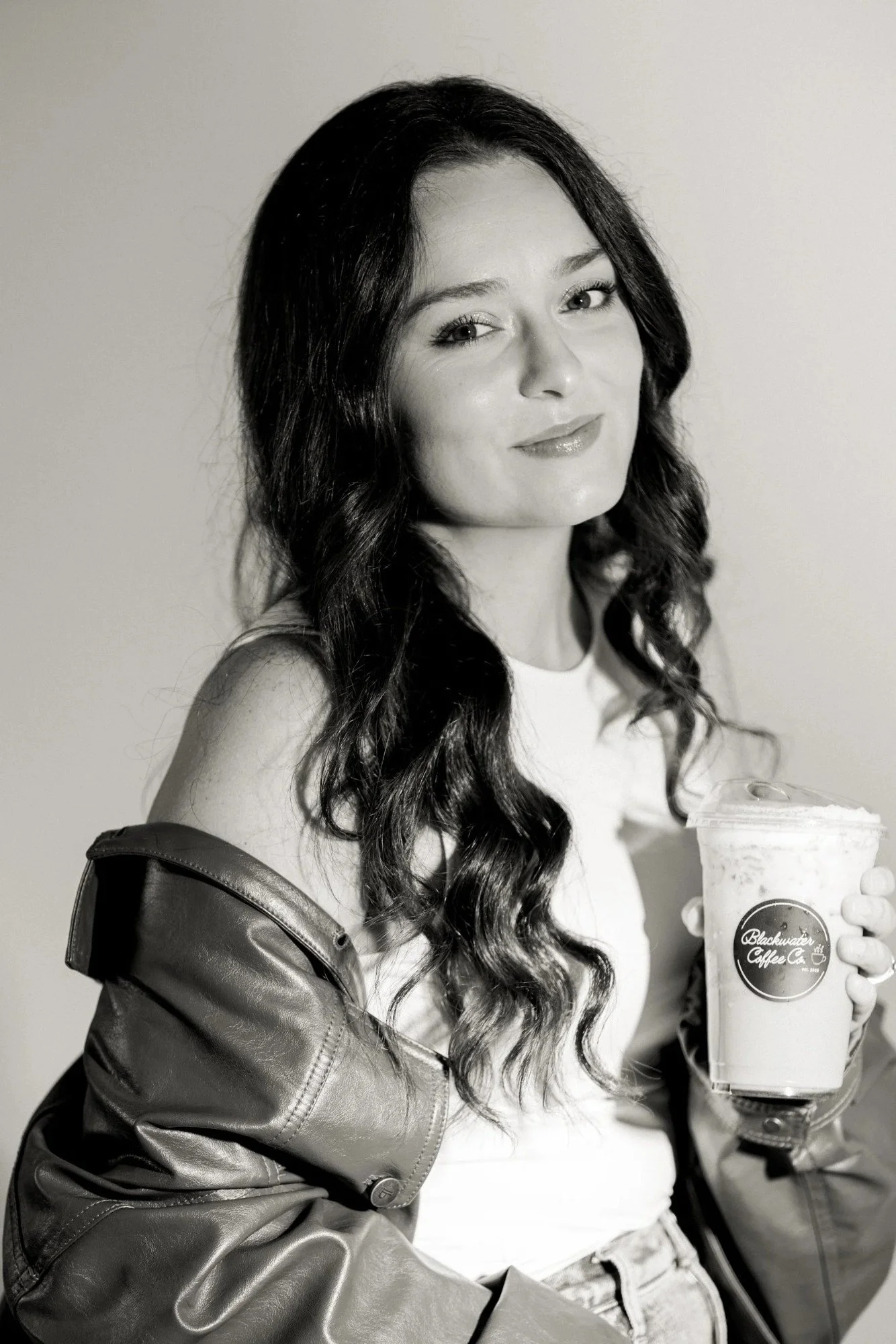 A woman with long wavy hair holding a cup from Blackwater Coffee Co. while sitting in a chair, wearing a white top and a shiny jacket, looking at the camera, in black and white.