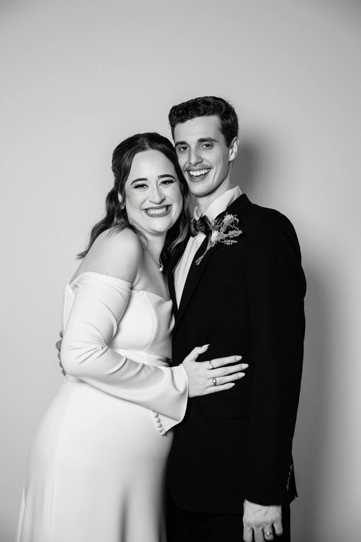 Black and white photo of a smiling bride and groom in wedding attire, embracing each other against a plain background.