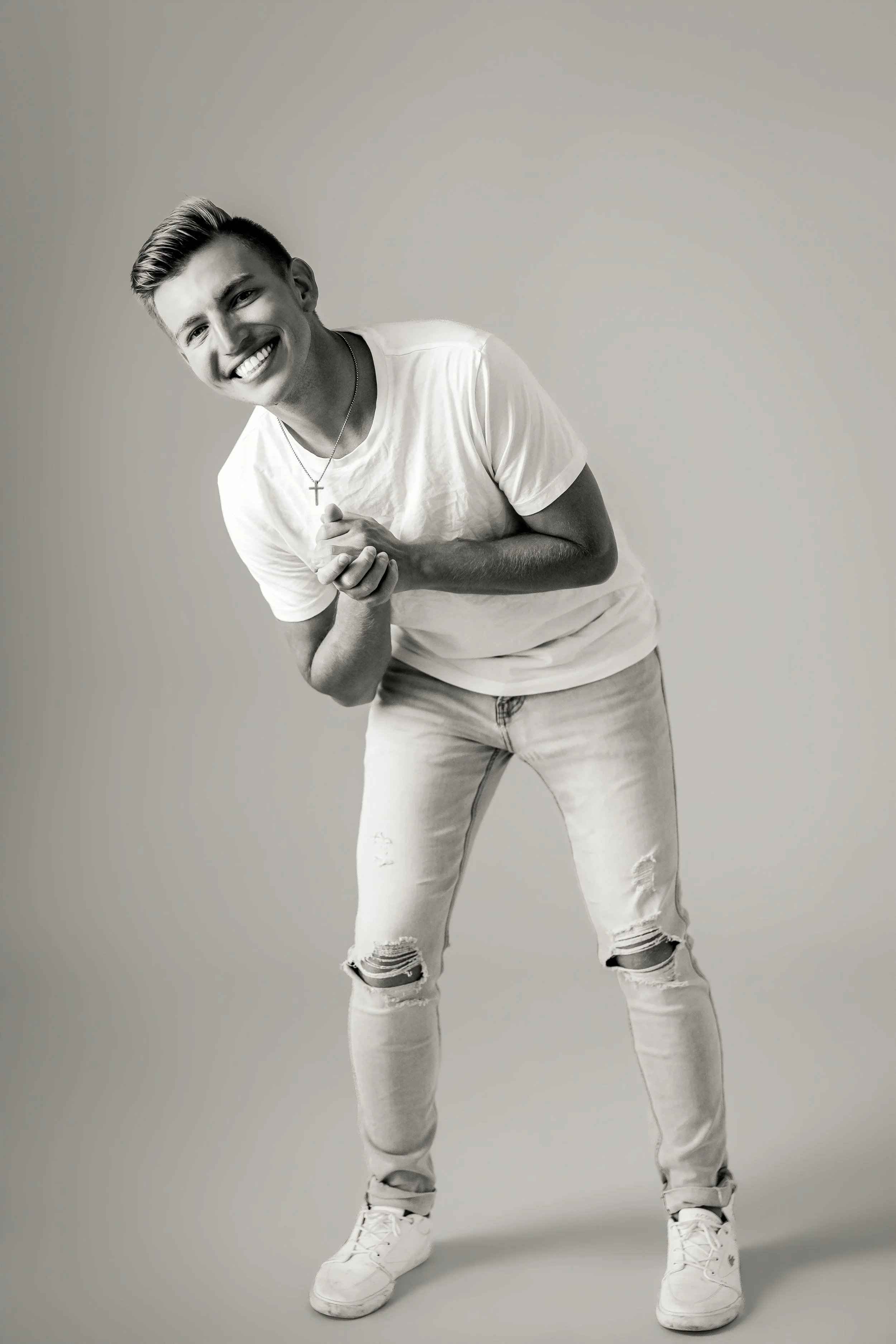 A young man with short hair, smiling, wearing a white t-shirt, ripped jeans, and sneakers, standing against a plain background, holding his hands together in front of him.