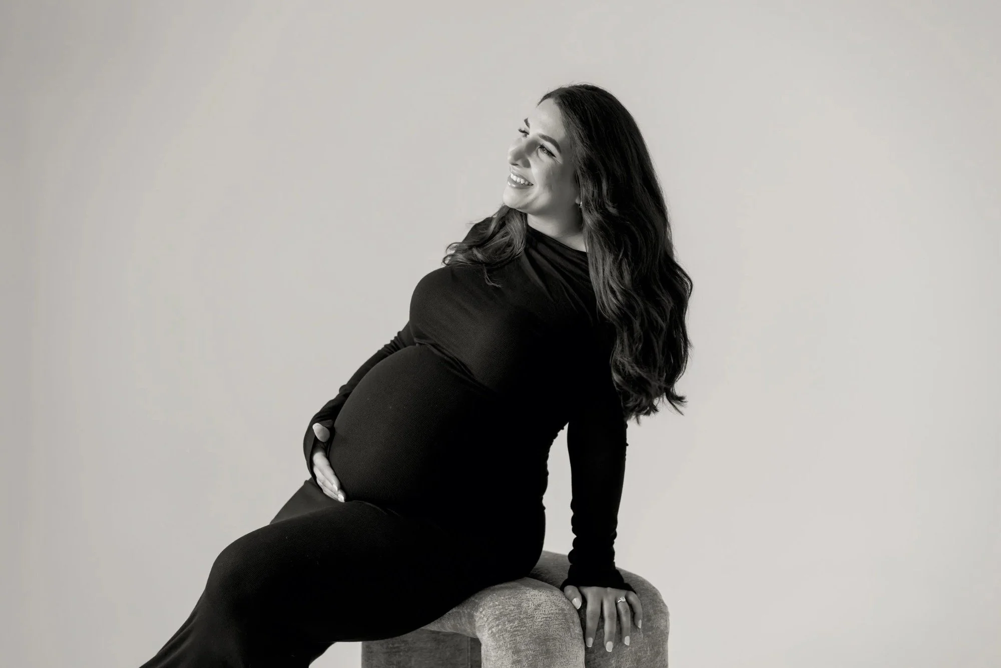 A woman with long dark hair wearing a black dress sitting on a cushioned surface, smiling and looking to her left, against a plain light-colored background.
