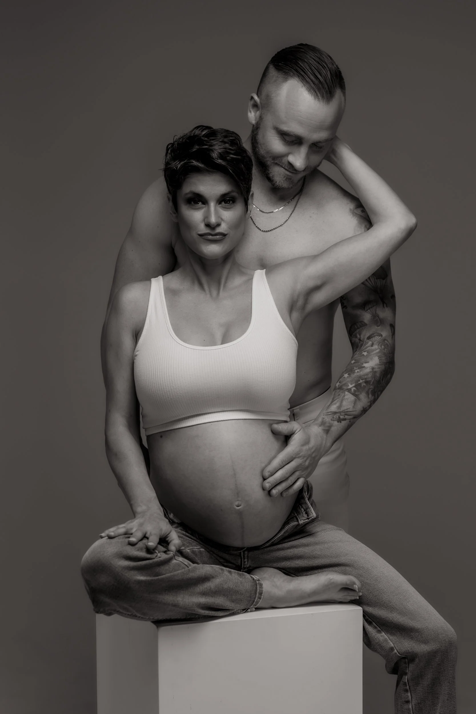Black and white photo of a woman with short hair, sitting cross-legged on a box, wearing a white tank top, pregnant, with a man behind her, both with calm expressions, the man with arm around the woman, both shirtless, with tattoos visible on the man