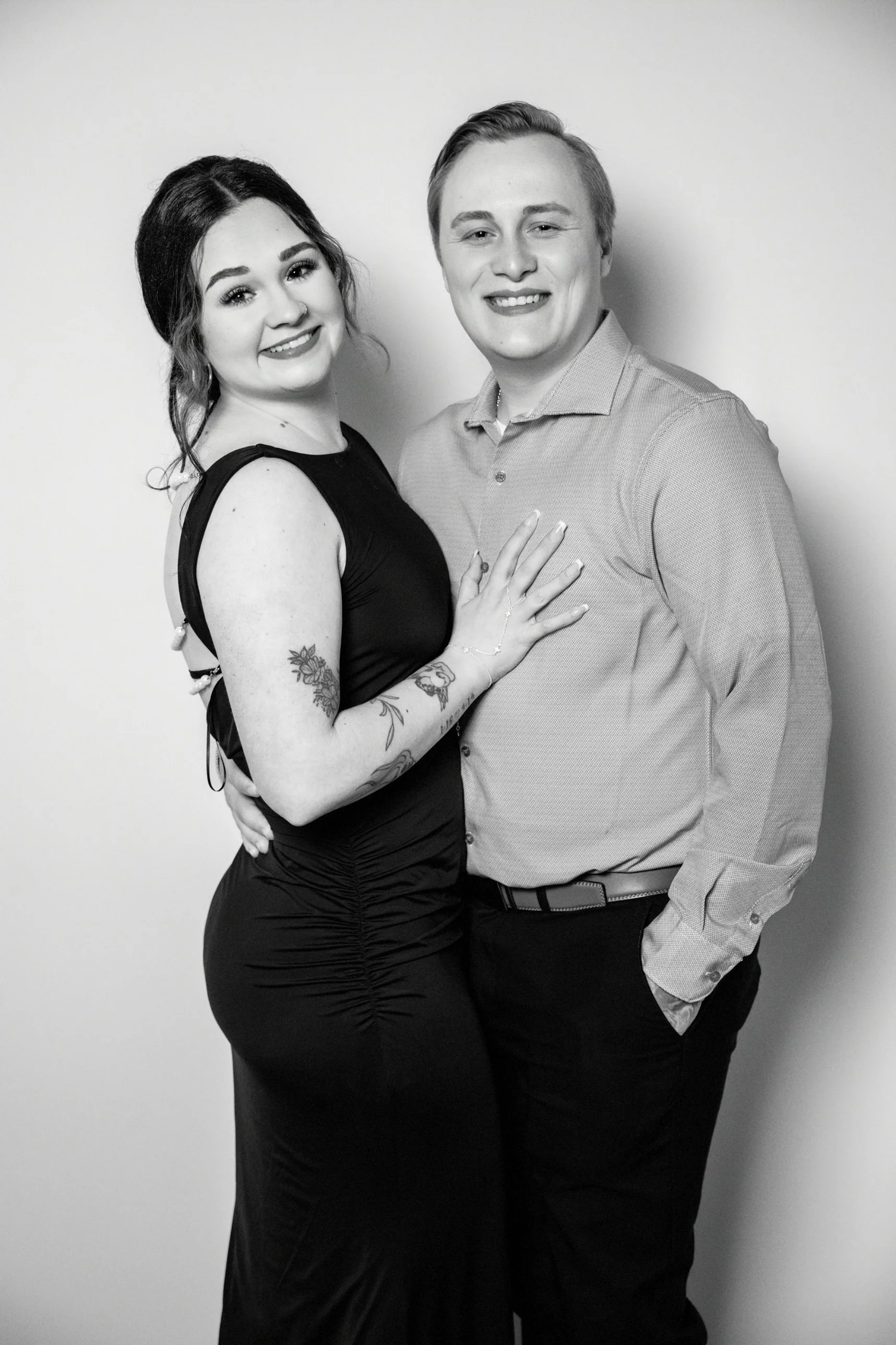 A black and white photo of a smiling couple standing closely together against a plain white wall. The woman has dark hair, visible tattoos on her upper arm, wearing a sleeveless dress, and is touching the man's chest. The man has light hair, is weari