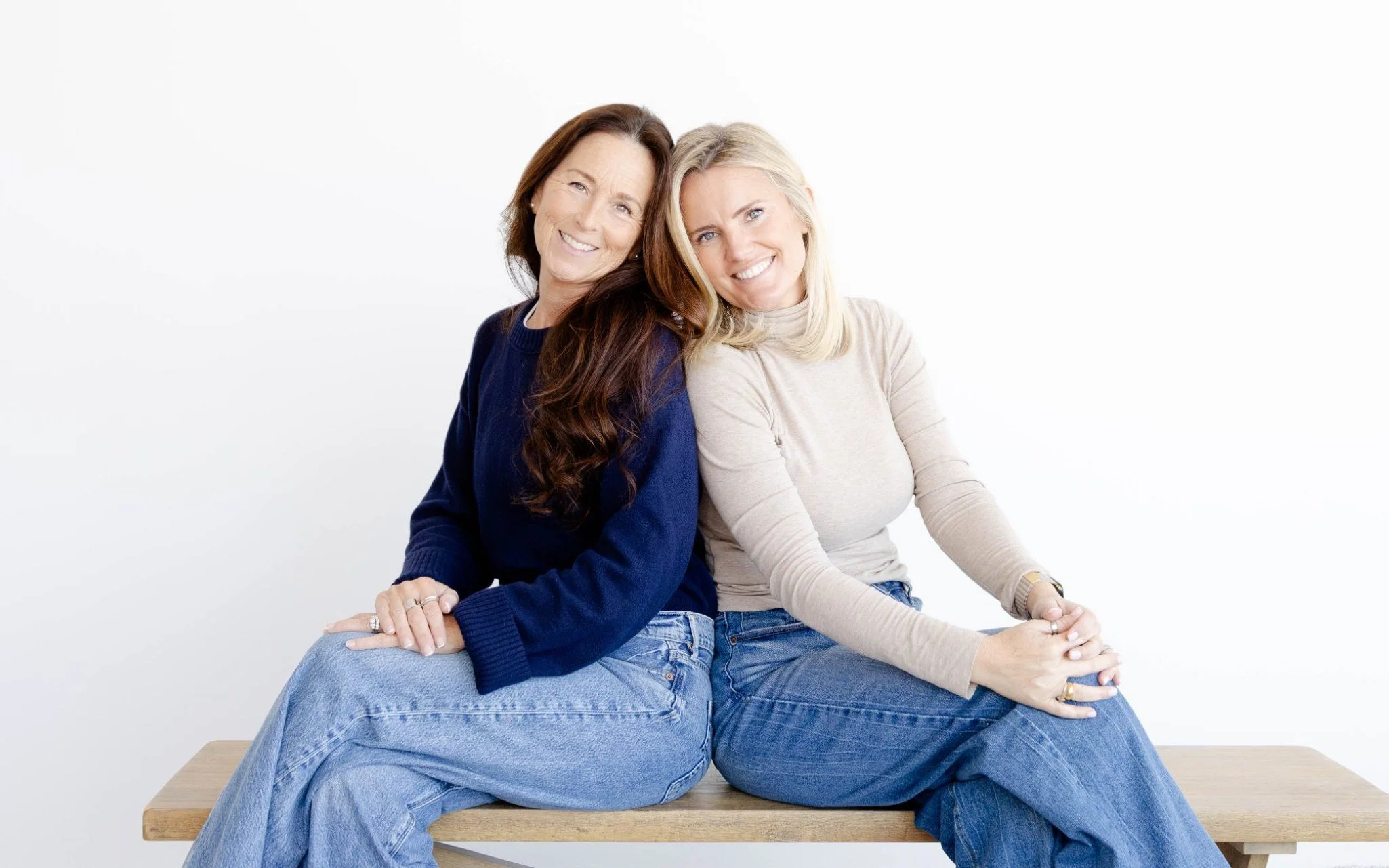 Two women sitting on a bench close together, smiling at the camera, against a plain white background.