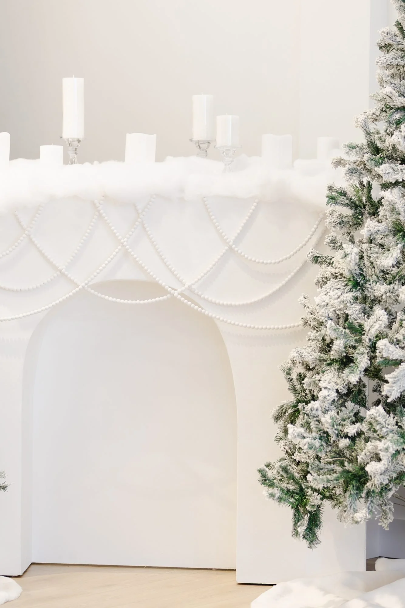 Decorative holiday scene featuring a white mantel with white candles and bead garlands, a snow-covered Christmas tree, and faux snow on the floor.