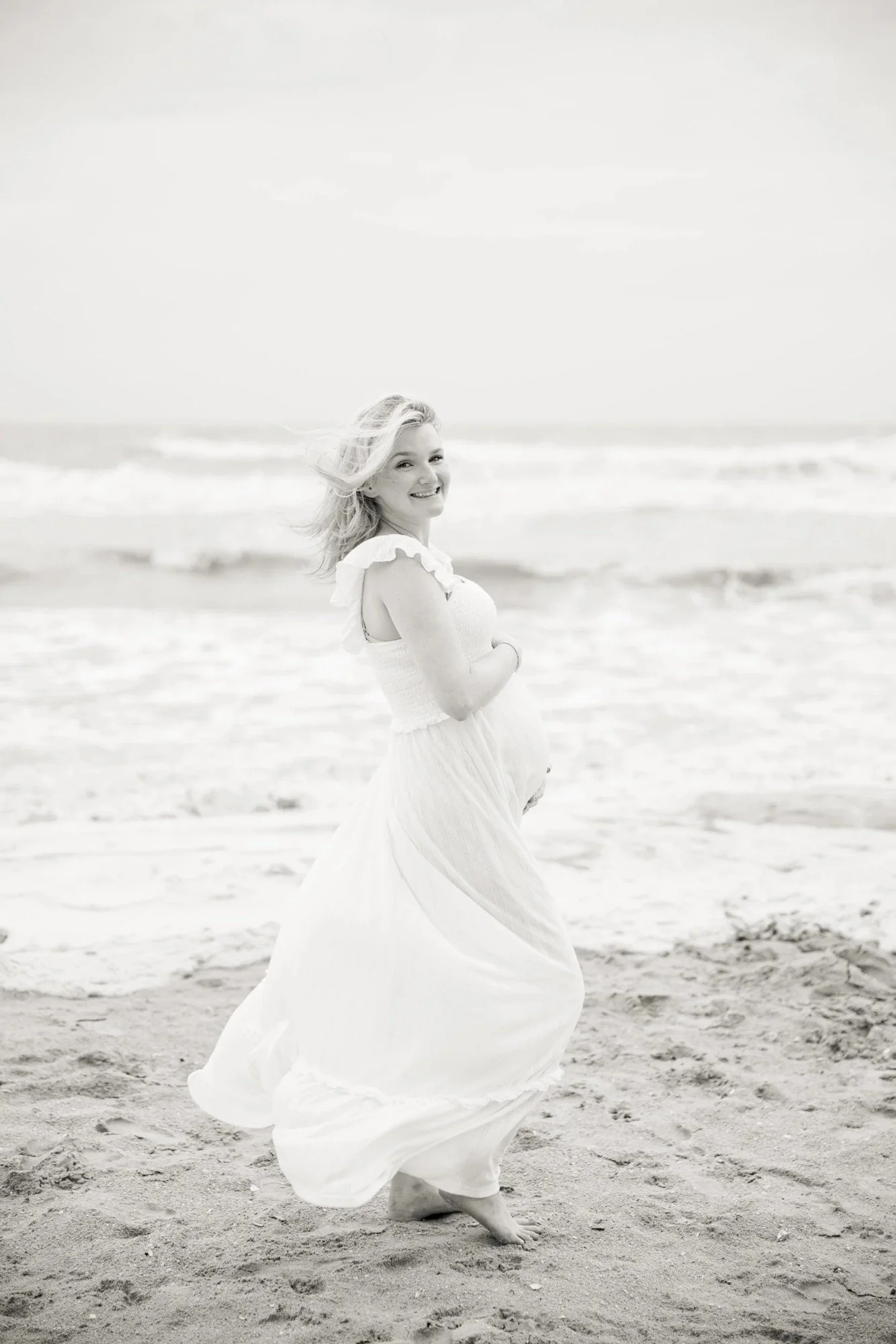 A pregnant woman in a white dress standing barefoot on the beach, smiling and looking over her shoulder, with ocean waves and cloudy sky in the background.