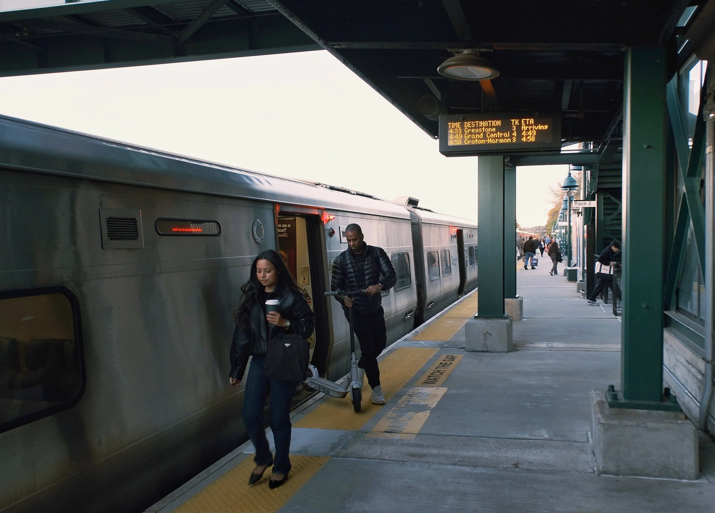 People boarding a subway train at an outdoor station with an electronic schedule display overhead.