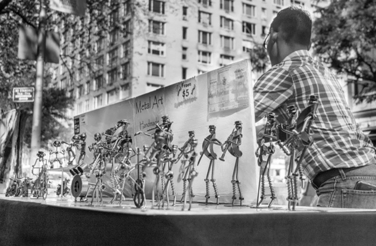 A man wearing glasses and a checkered shirt is selling metal art sculptures of musicians at an outdoor market. The sculptures are displayed on a table with a sign that reads "Metal Art Handcrafted from $5." High-rise buildings and trees are visible i