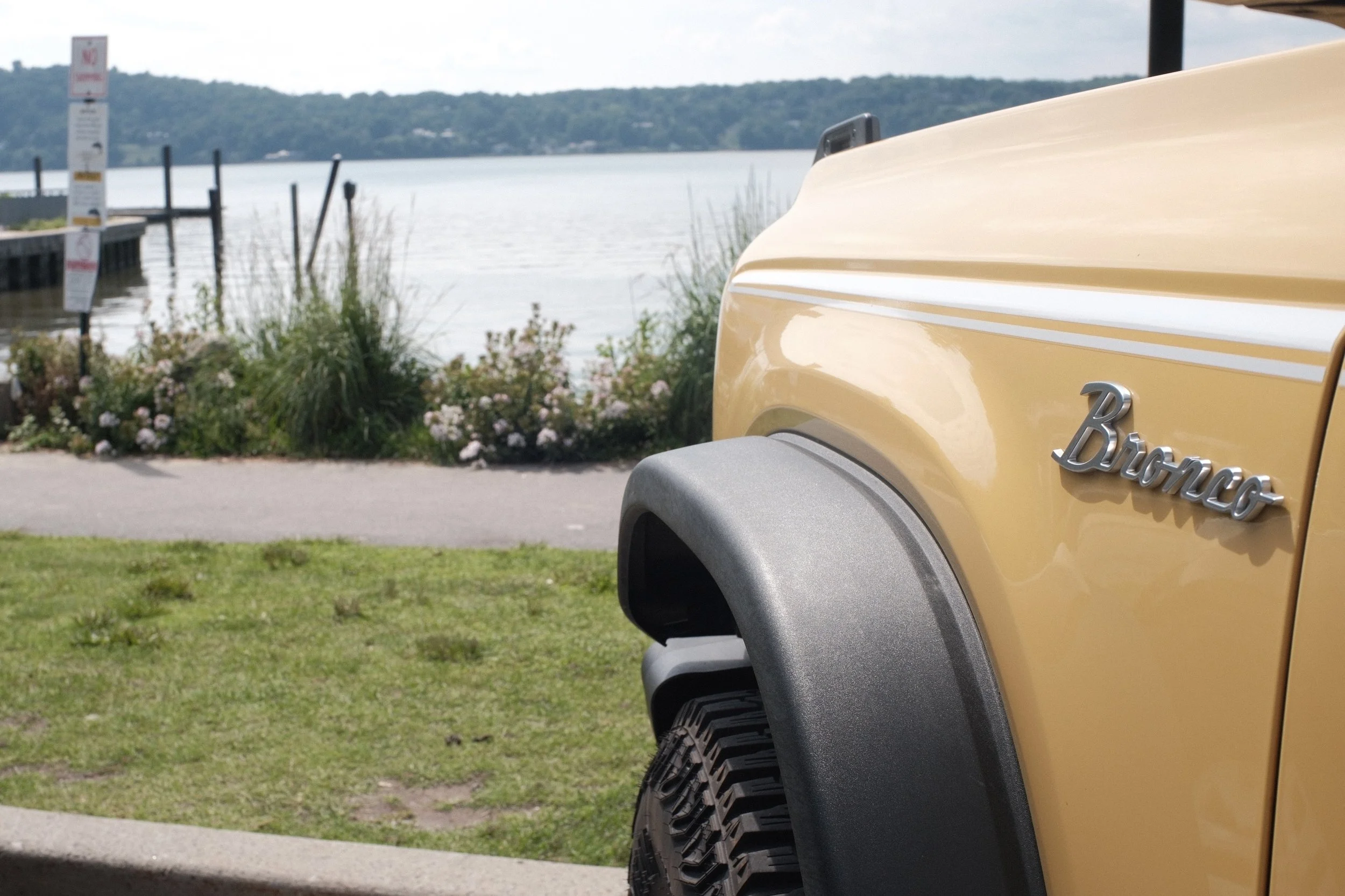 Close-up of the front side of a yellow GMC Hummer electric vehicle parked near a lakeside with a dock, water, and hills in the background.