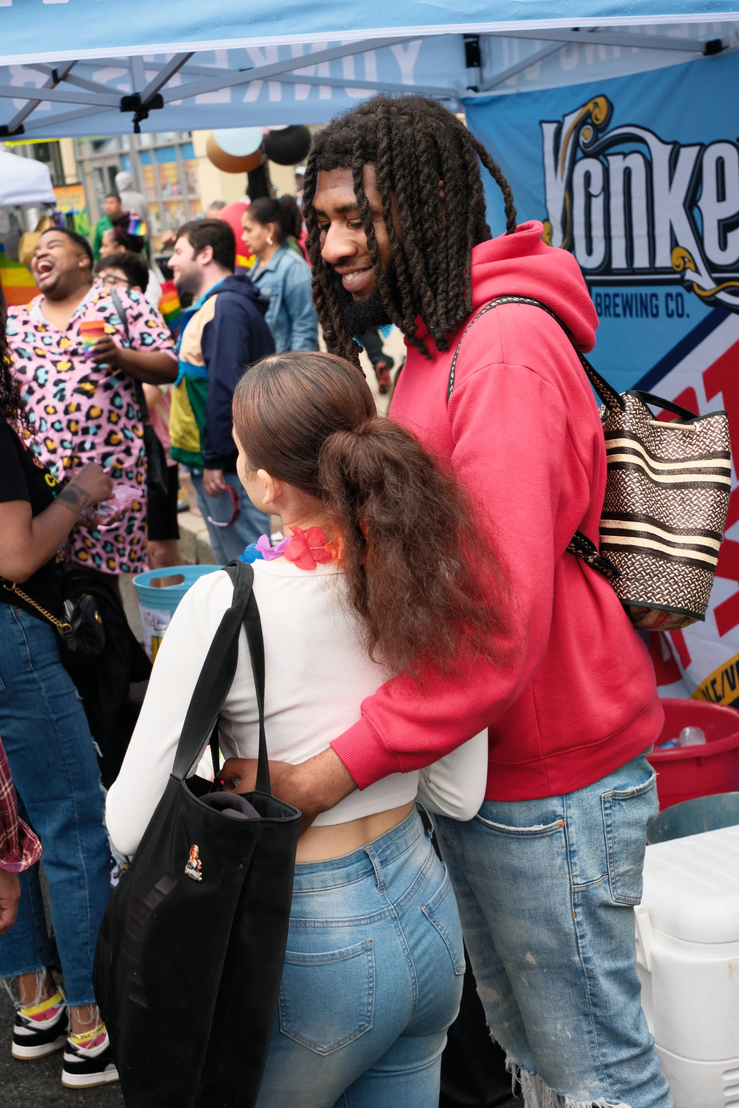 A group of people at an outdoor event, with a man in a red hoodie and dreadlocks near a woman in a white top and curly hair, both standing close together. In the background, others are laughing and talking, and there is a blue banner with the word 'Y