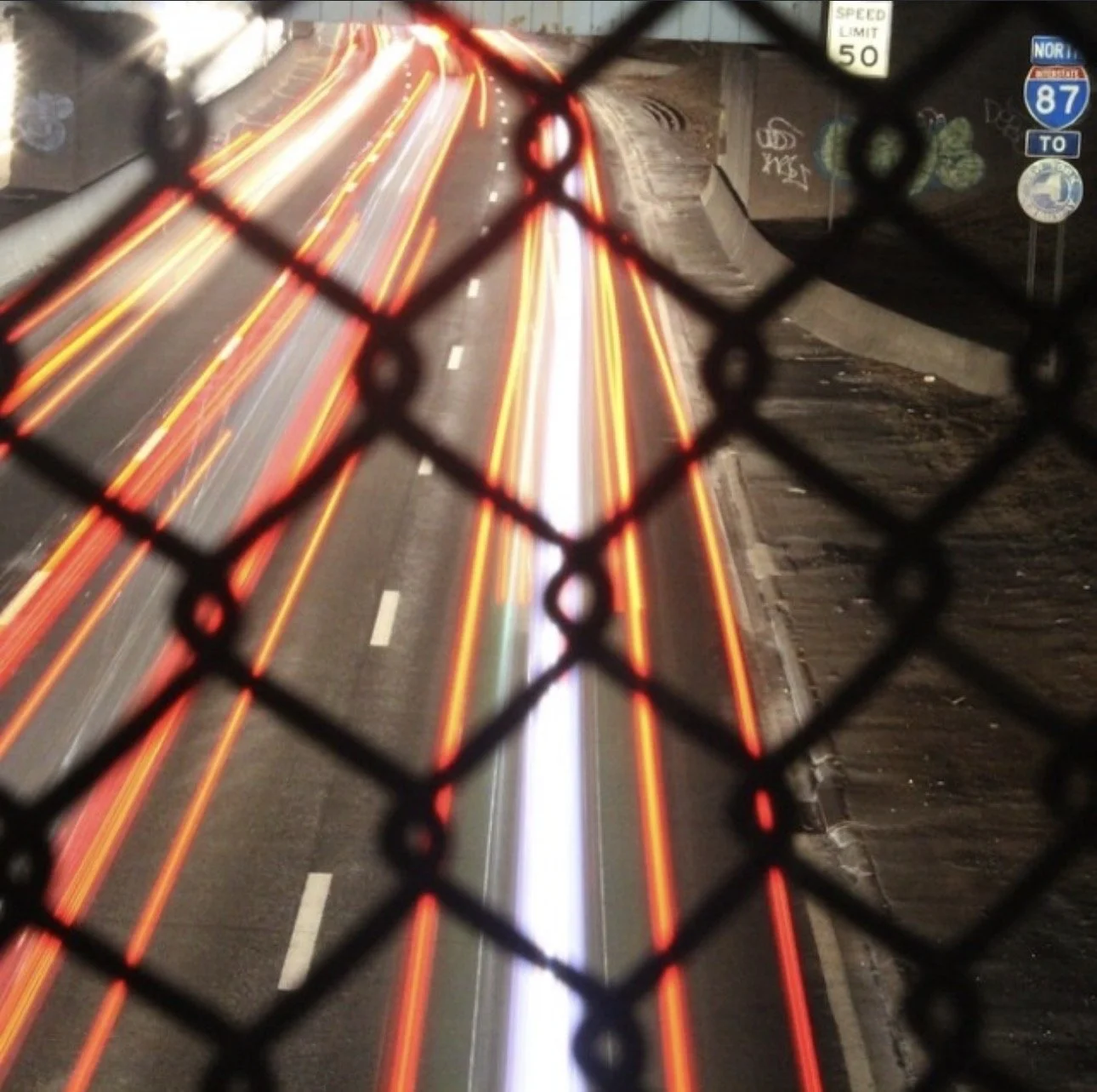 Long exposure photo of a highway with red and white light trails from vehicles taken at night, viewed through a chain-link fence.