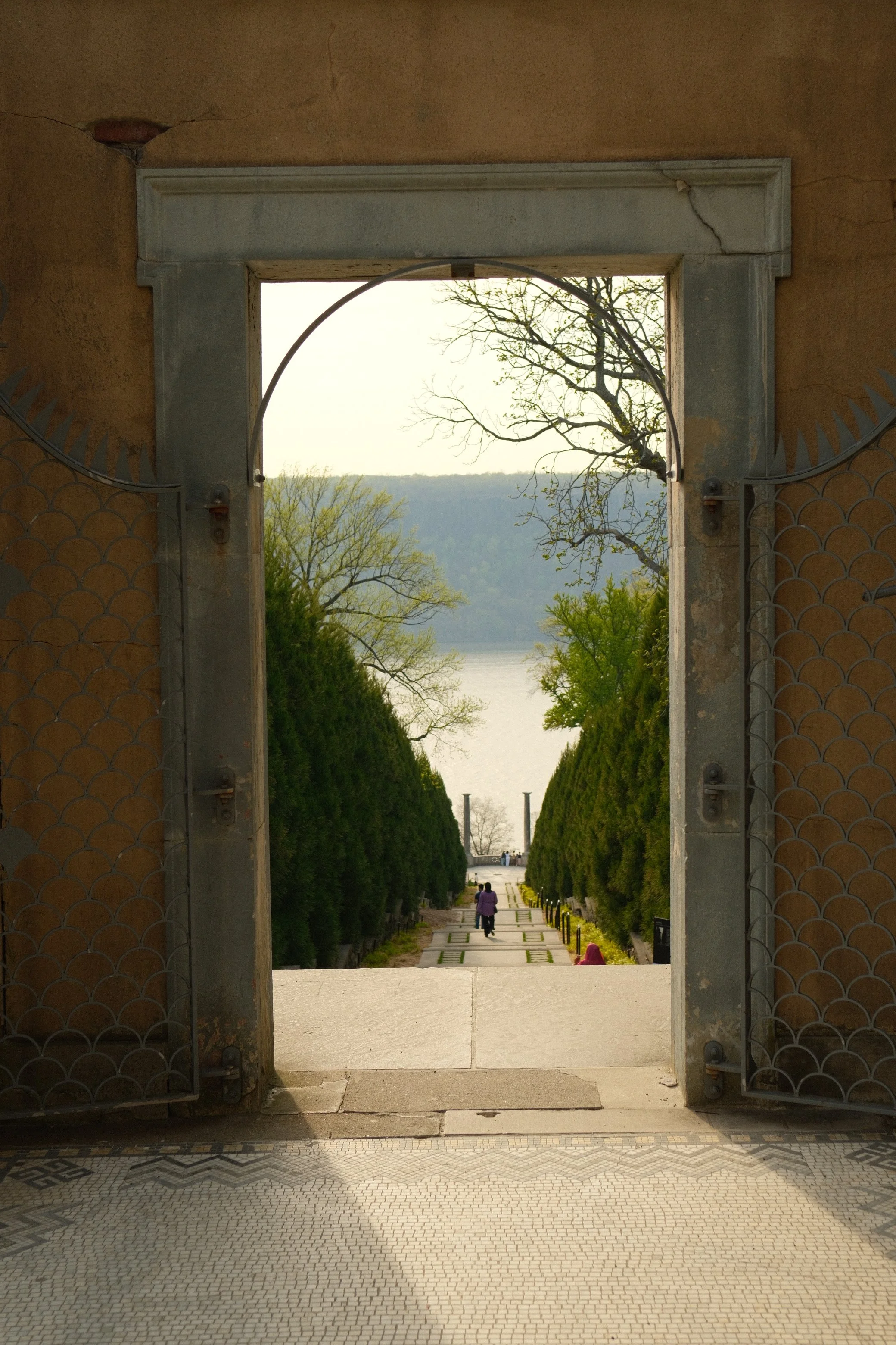 Open metal gates frame a view of a paved walkway lined with tall green trees and a lake in the background. People walk along the path, and there's a person sitting on the ground to the right.