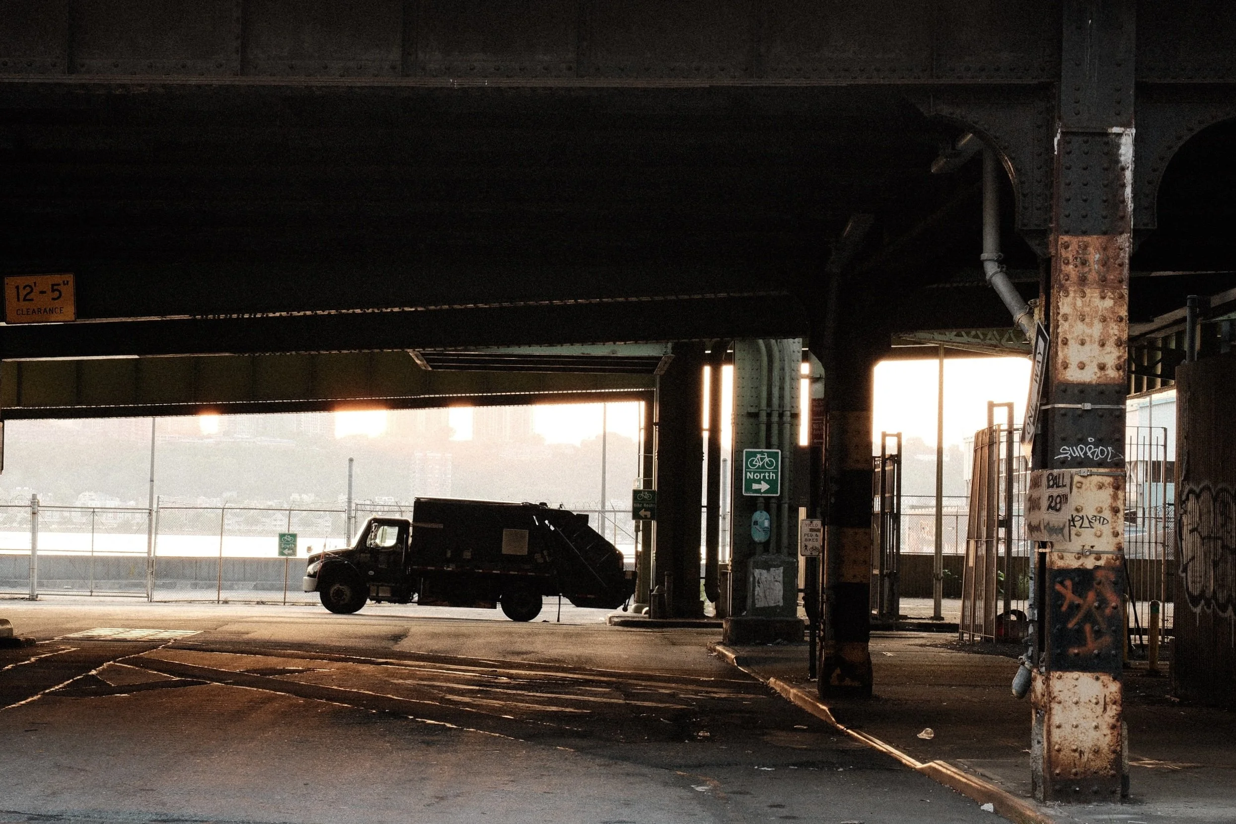 A parking lot under a bridge at sunset, with a black vehicle parked in the foreground. The area is fenced and has various small signs, including a green bicycle route sign indicating north. Graffiti is visible on a rusted pillar.