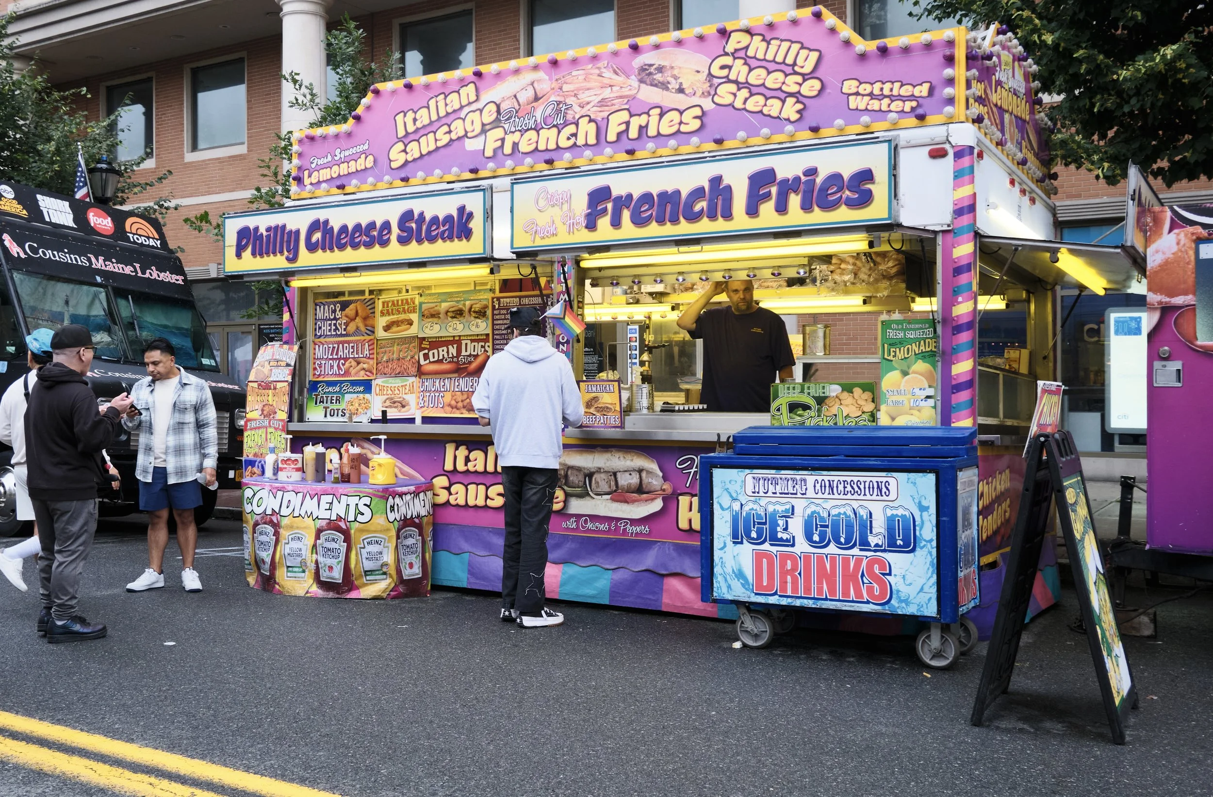A colorful outdoor food stall selling French fries, cheesesteaks, sausage, and lemonade. Several people are gathered around the stall, which has bright signage and menus.