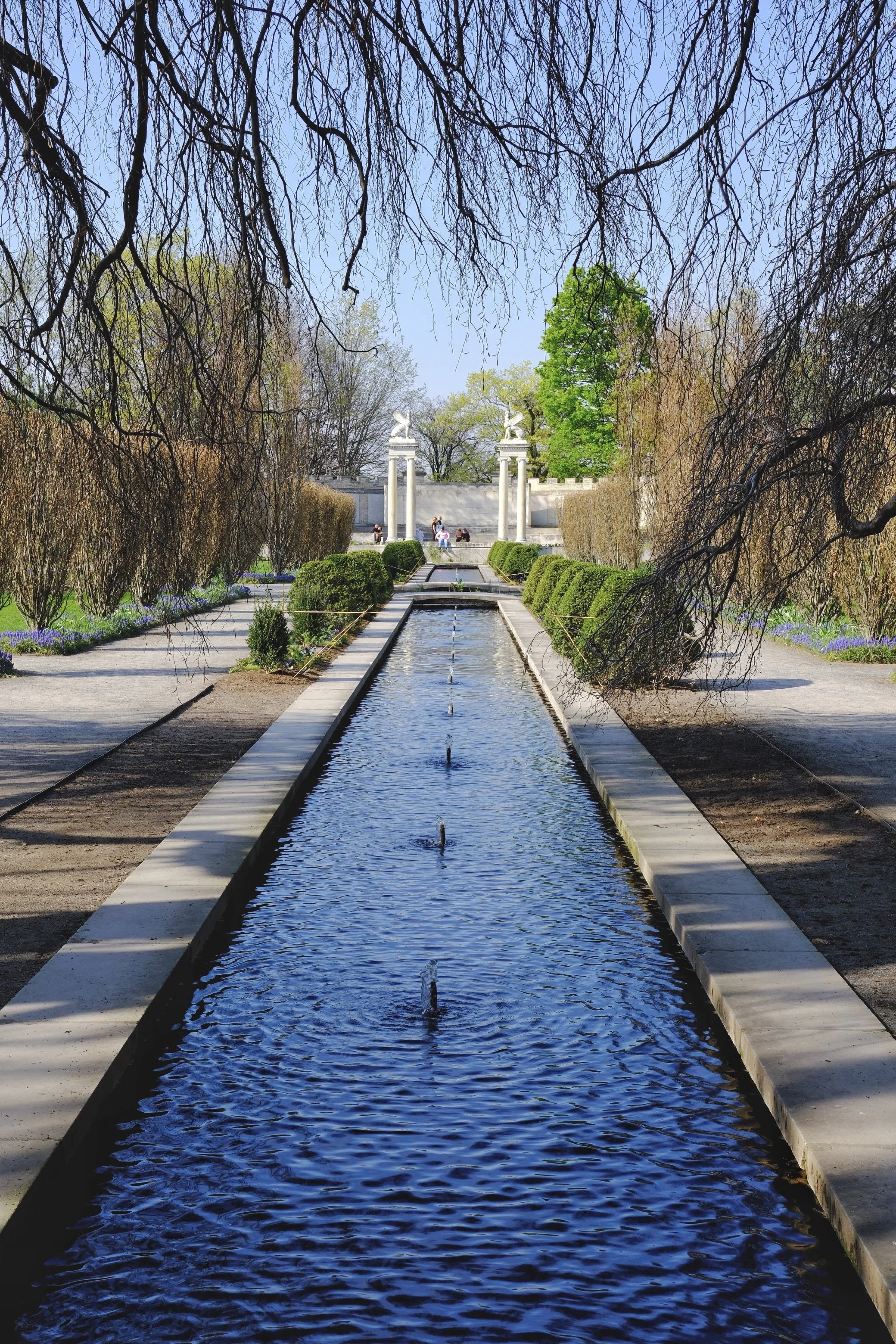 A long rectangular water feature in a garden, with water jets, green bushes, and trees around it, leading towards a classical white sculpture with winged figures on columns, framed by leafless branches at the top.