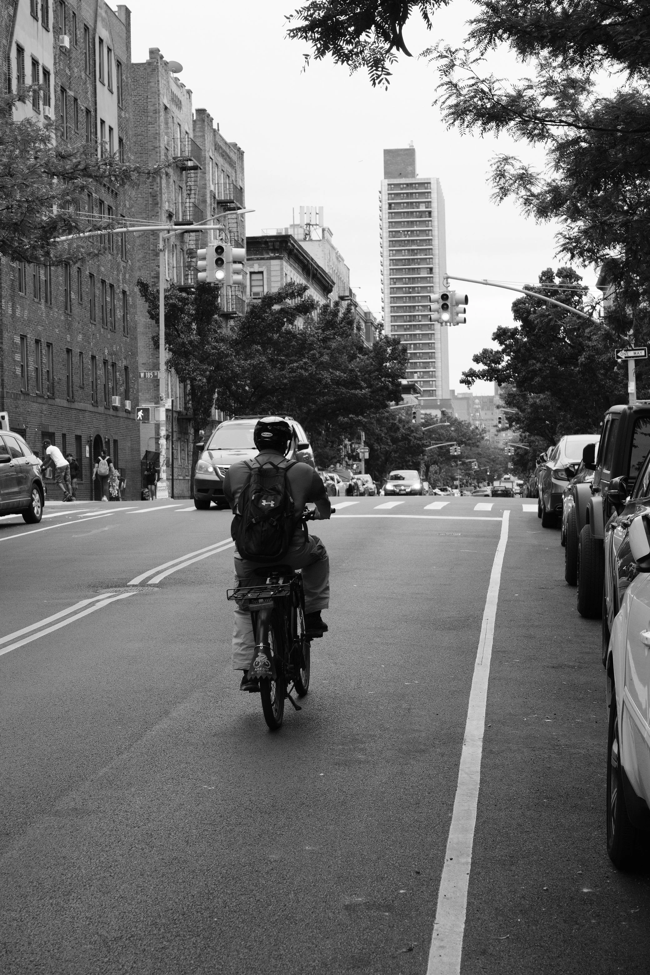 Black and white photo of a city street with cars, a man riding a bicycle, traffic lights, buildings, and trees.