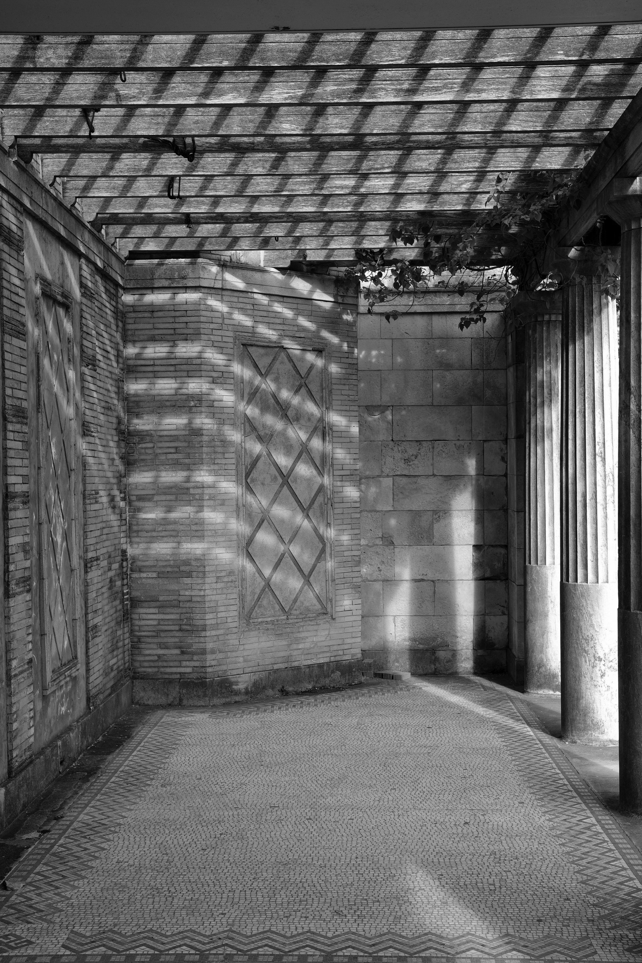Black and white photo of a building corner with brick and stone walls, decorative doors, column, trellis with climbing plants, and a patterned stone walkway.