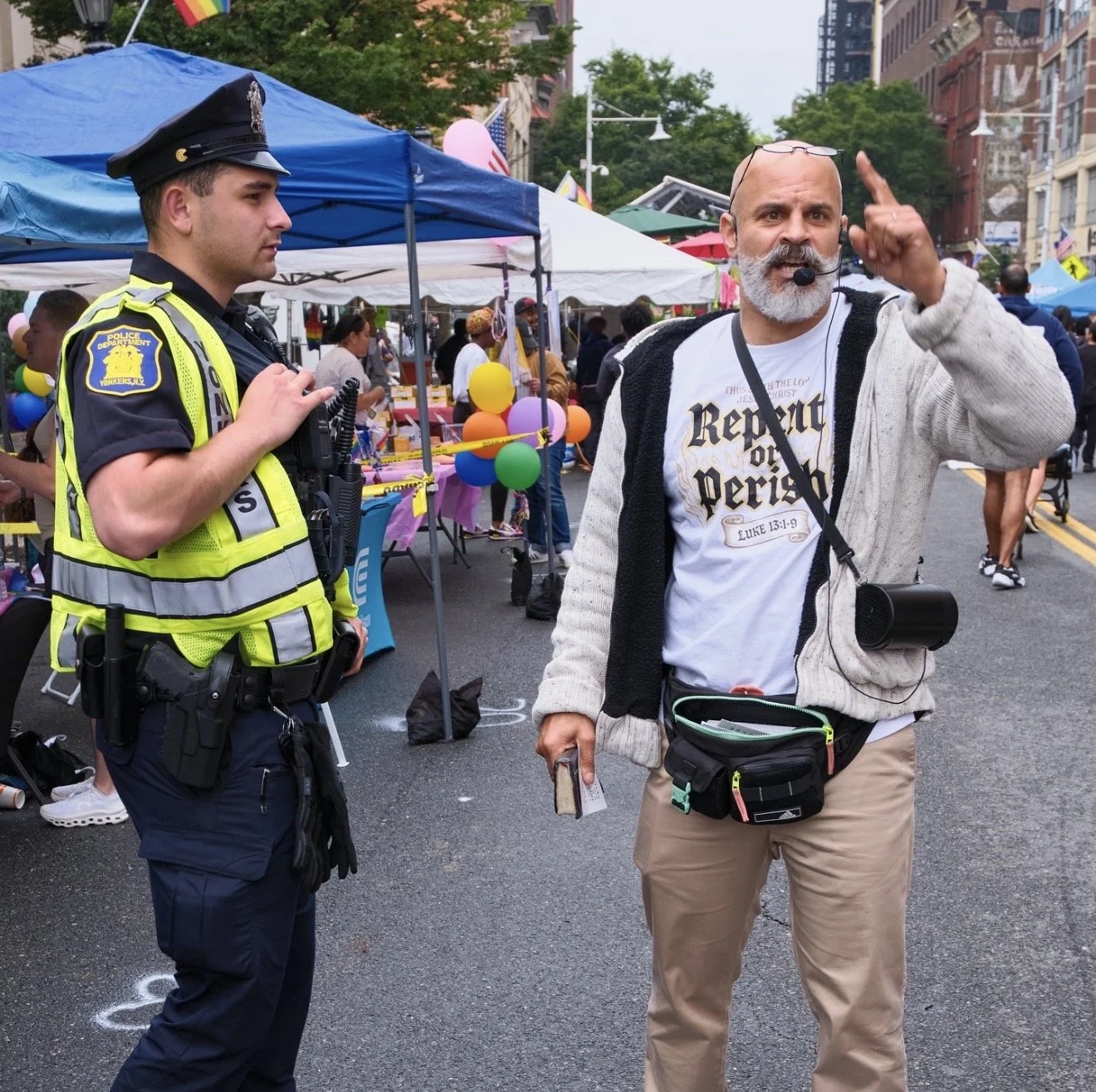 A man with glasses and a beard wearing a microphone headset gestures with his finger while talking to a police officer at a street fair. The police officer is dressed in a navy uniform with a yellow reflective vest, and stands under a blue tent with 