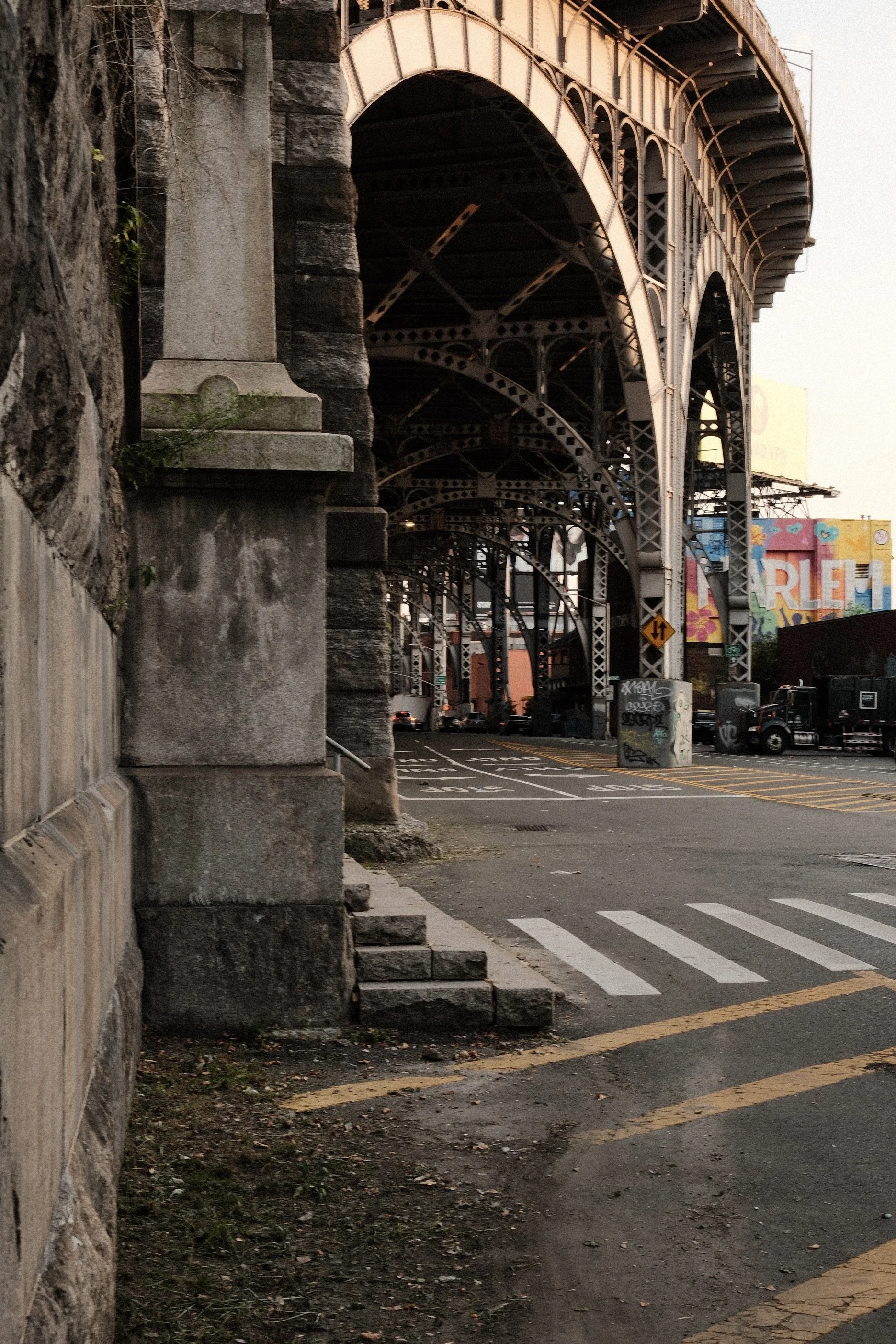 View of the underside of a large iron bridge spanning over a street in an urban area, with graffiti-covered walls and cars parked below, seen during the evening or early dawn.