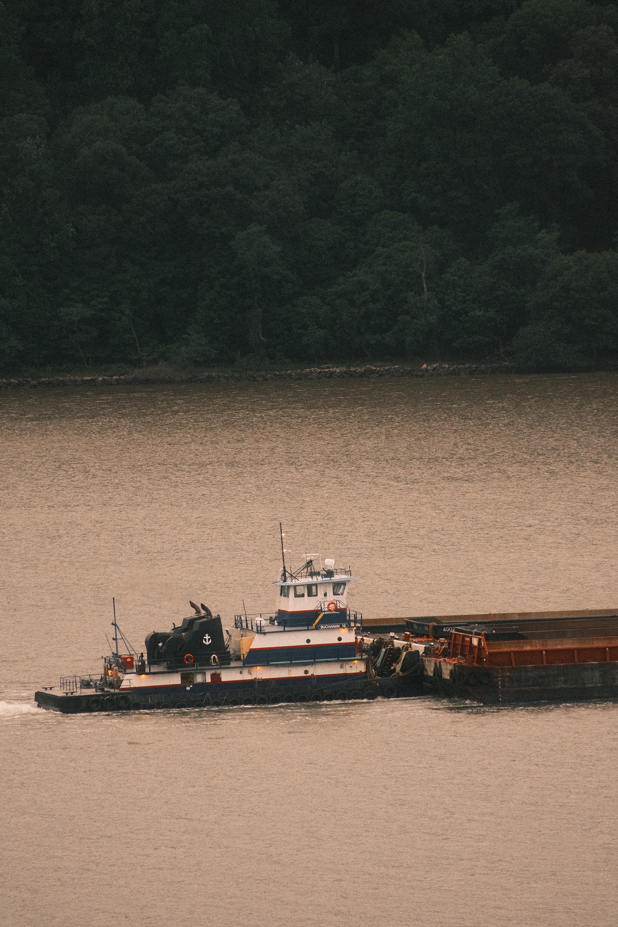A tugboat moving across a body of water with a wooded shoreline in the background.