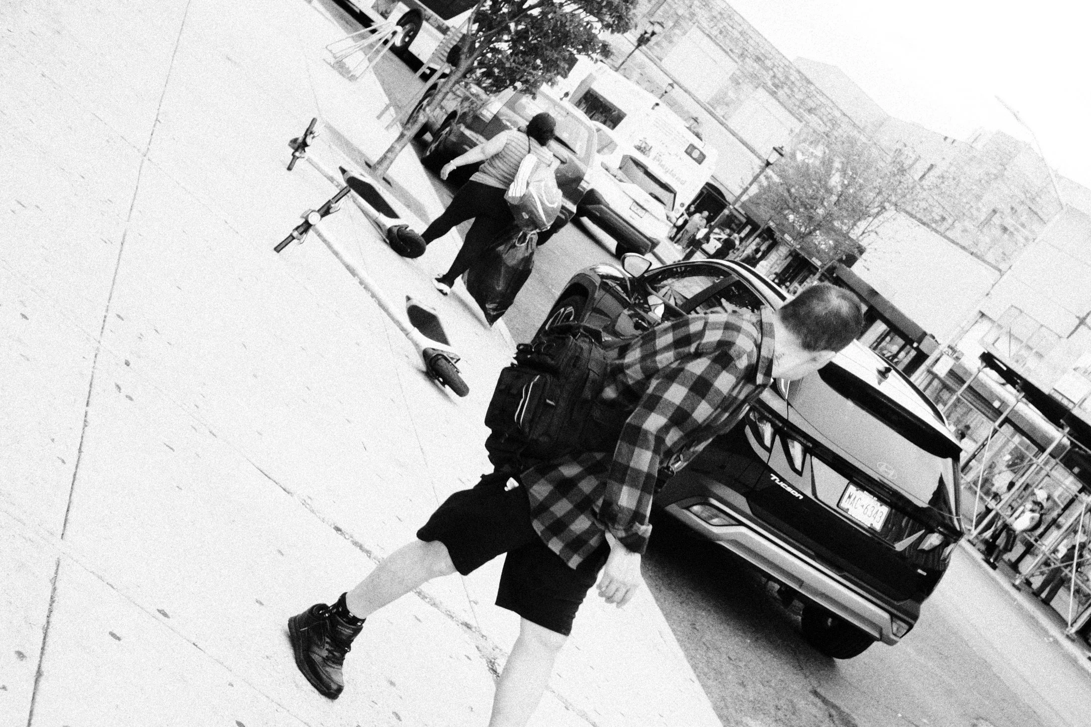 A black-and-white photo of a woman crossing a city street at a crosswalk, carrying a backpack and shopping bags, dressed in a striped shirt and dark pants. There are parked cars, a fire hydrant, and a storefront in the background.