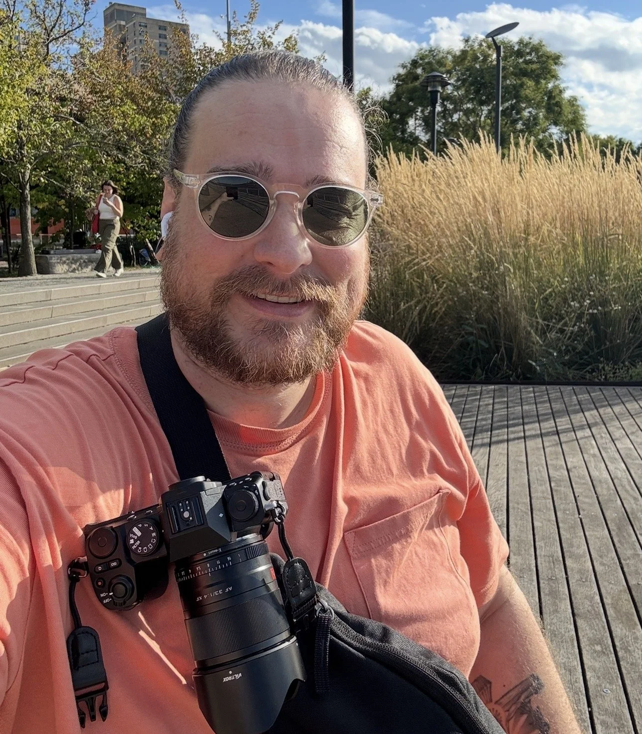 A smiling man with glasses and a beard taking a selfie outdoors on a sunny day, wearing a coral-colored shirt with a camera hanging around his neck. In the background, there's a woman walking near trees and urban buildings, with tall grasses and a partly cloudy sky.
