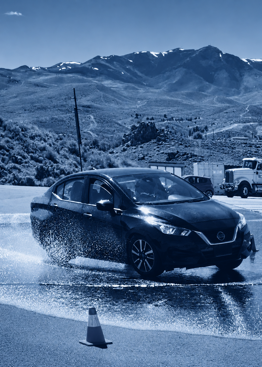 Black Nissan sedan driving through water spray in a mountainous area with snow-capped peaks in the background, orange traffic cone in foreground, and trucks parked nearby.