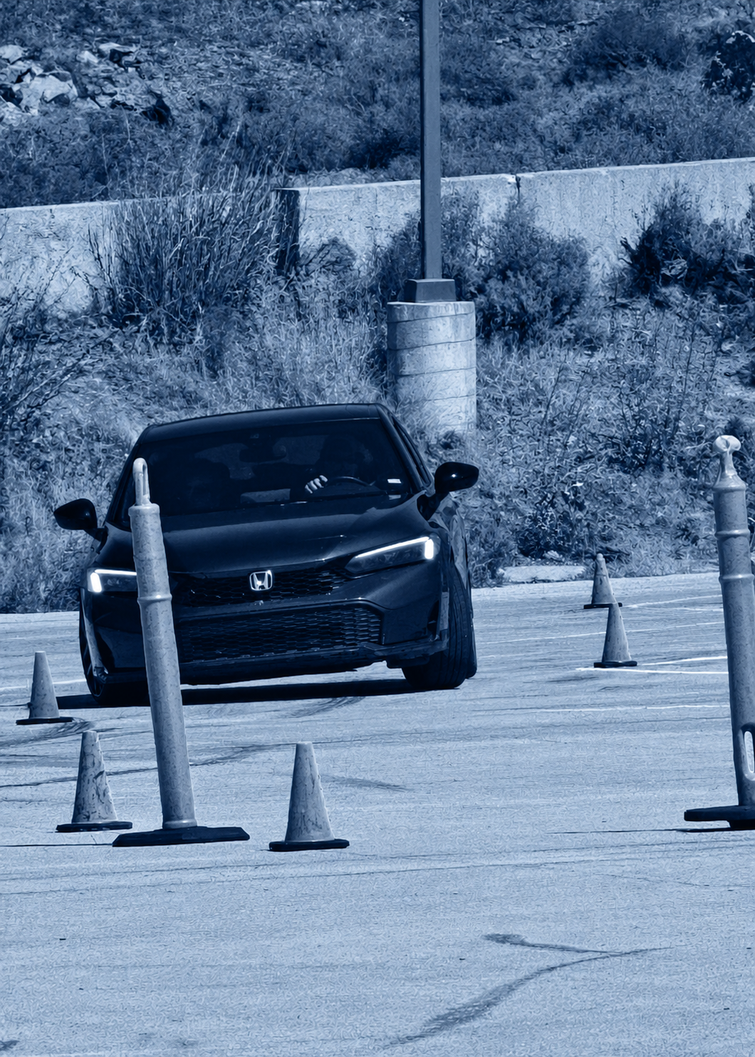 A black Honda car parked in a parking lot with orange cones around it and a backdrop of bushes and a stone wall, in a monochrome blue filter.