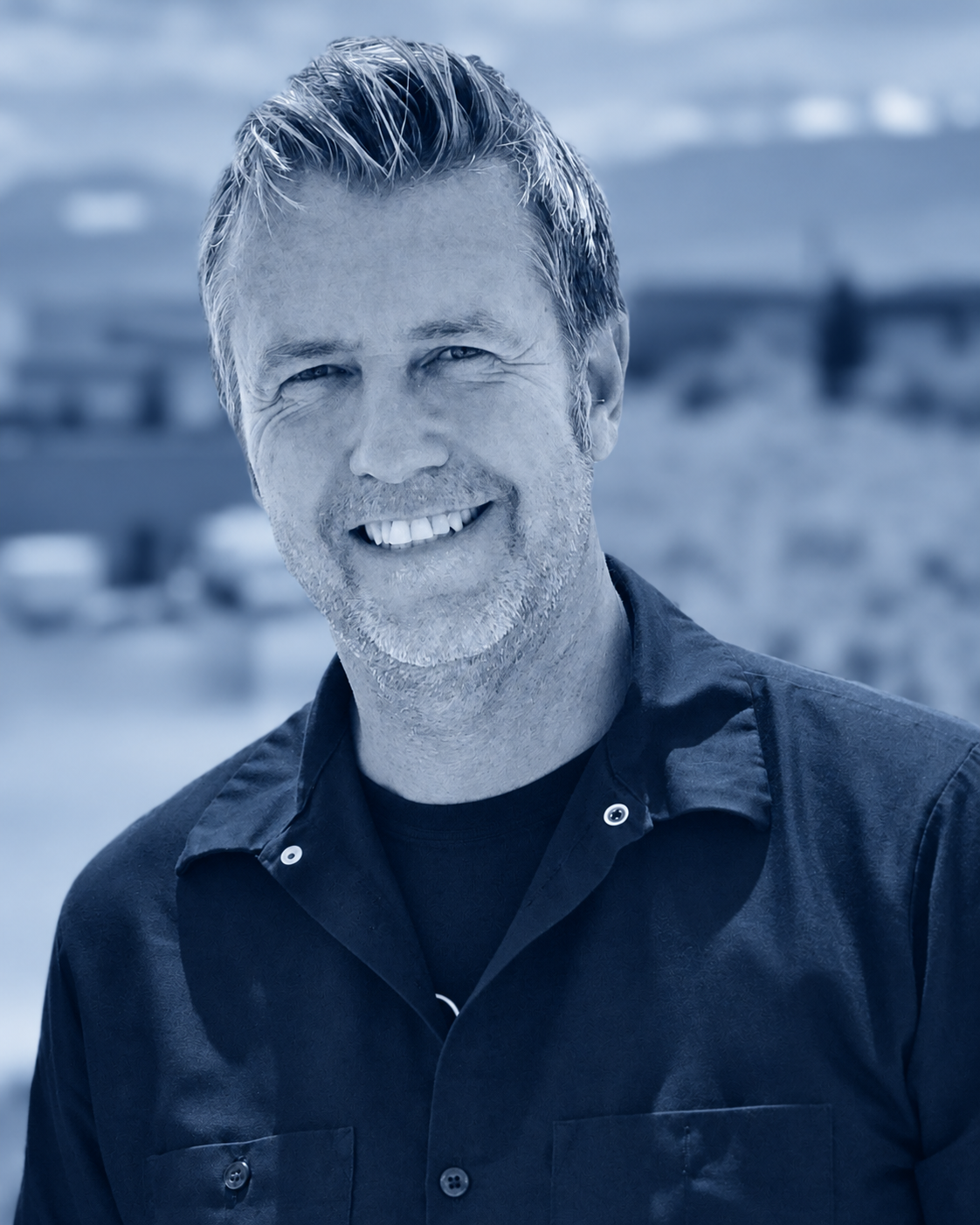 A smiling man with salt-and-pepper hair and beard, wearing a dark shirt, standing outdoors with blurred background.