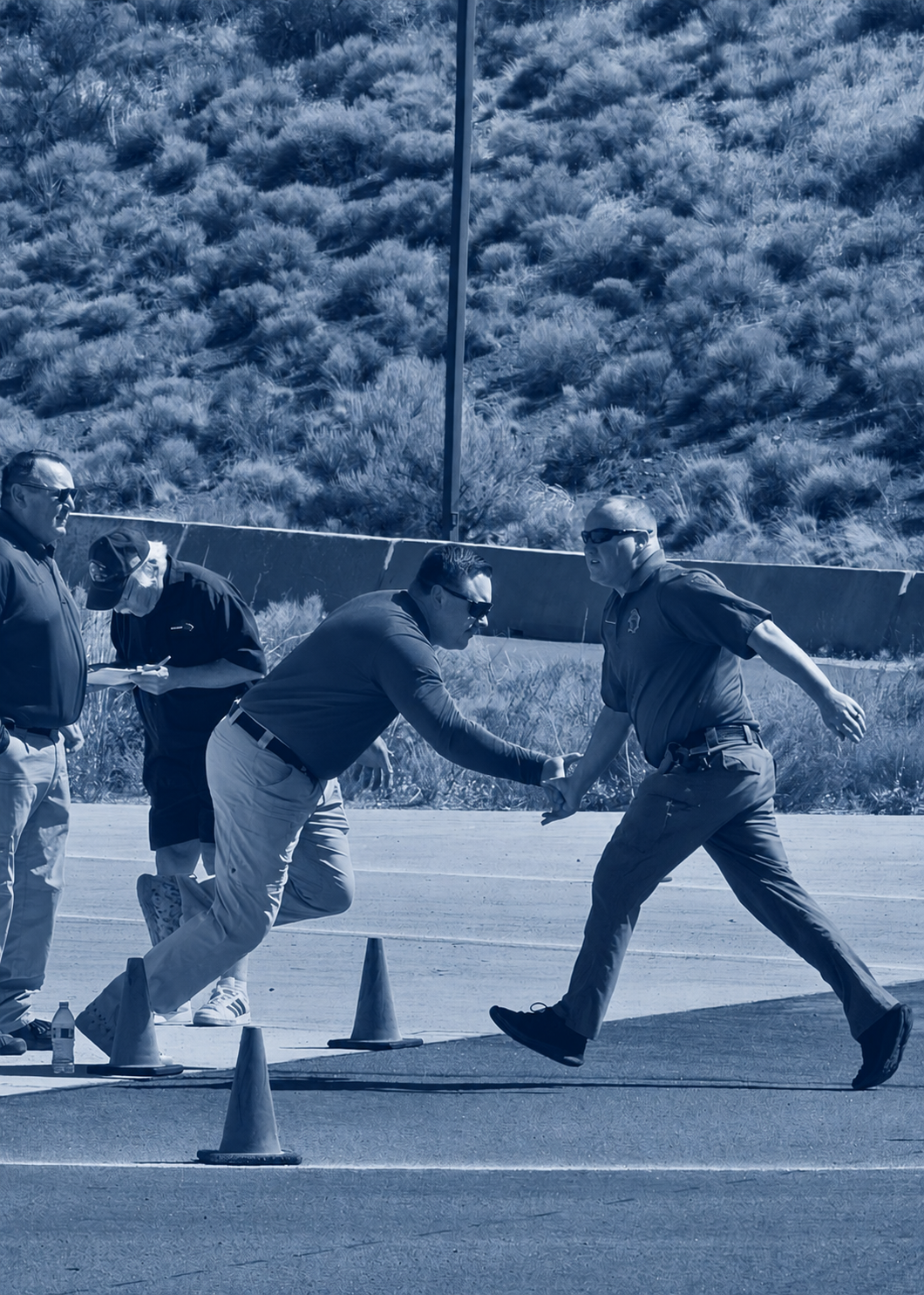 Two men shaking hands during a race or event, with several officials or volunteers nearby, marking a finishing line with traffic cones on an outdoor road surrounded by desert plants.