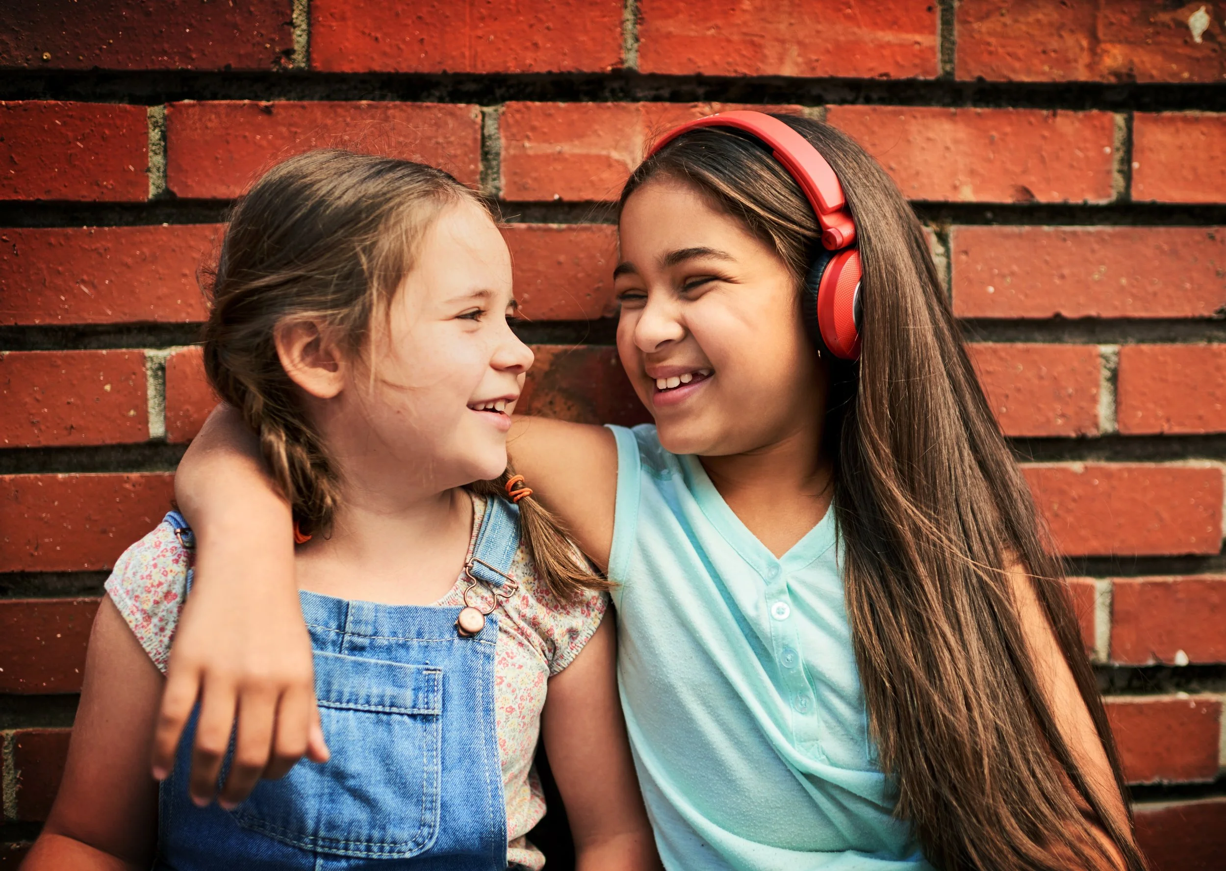 Two smiling girls hug in front of a brick wall, with one wearing red headphones.