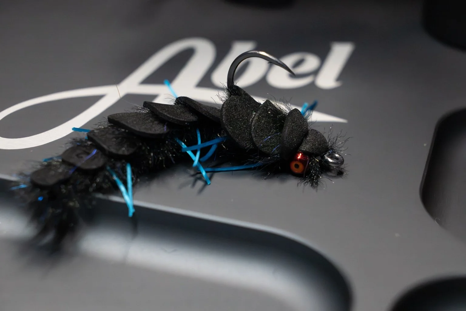 Long black fly with overlapping foam-back segments, blue legs and orange eyes.