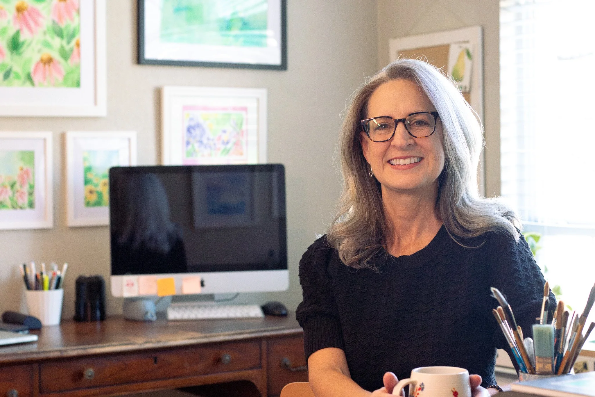 Smiling woman (Amelia Audish) with glasses holding a coffee mug in a home art studio with framed artwork and paintbrushes