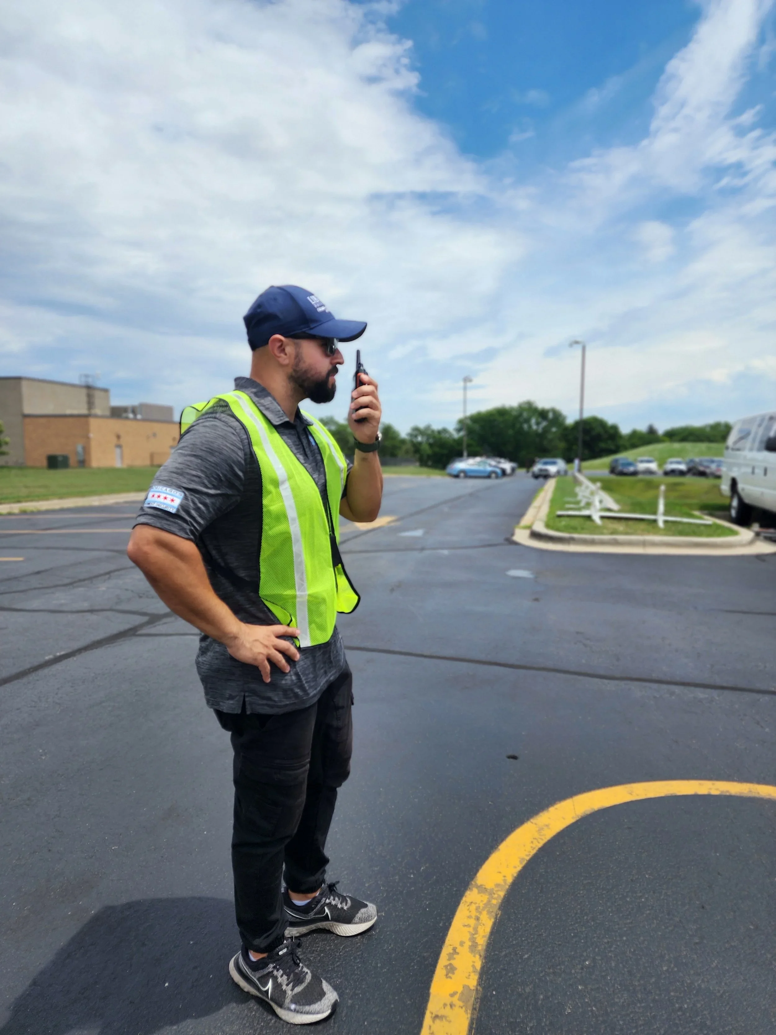 A man wearing a blue cap, sunglasses, a gray shirt with a police patch on the sleeve, and a bright yellow safety vest is standing in a parking lot, holding a two-way radio to his mouth, with his left hand on his hip. The parking lot has several cars, a grassy area with trees, and a building in the background. The sky is partly cloudy.