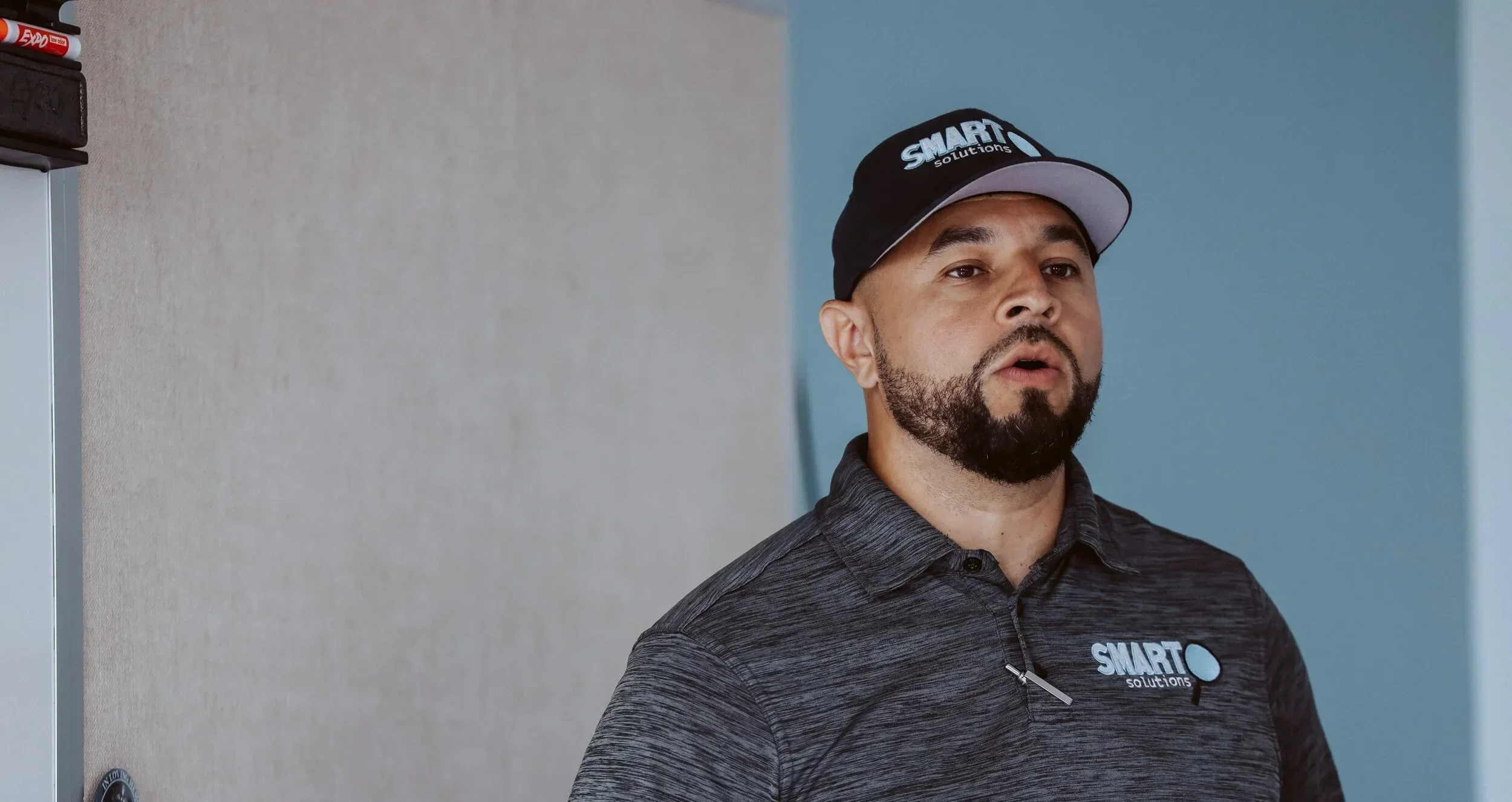 A man wearing a black baseball cap and dark gray polo shirt with 'SMART Solutions' logo, standing indoors against a blue and gray wall, looking slightly to his right.