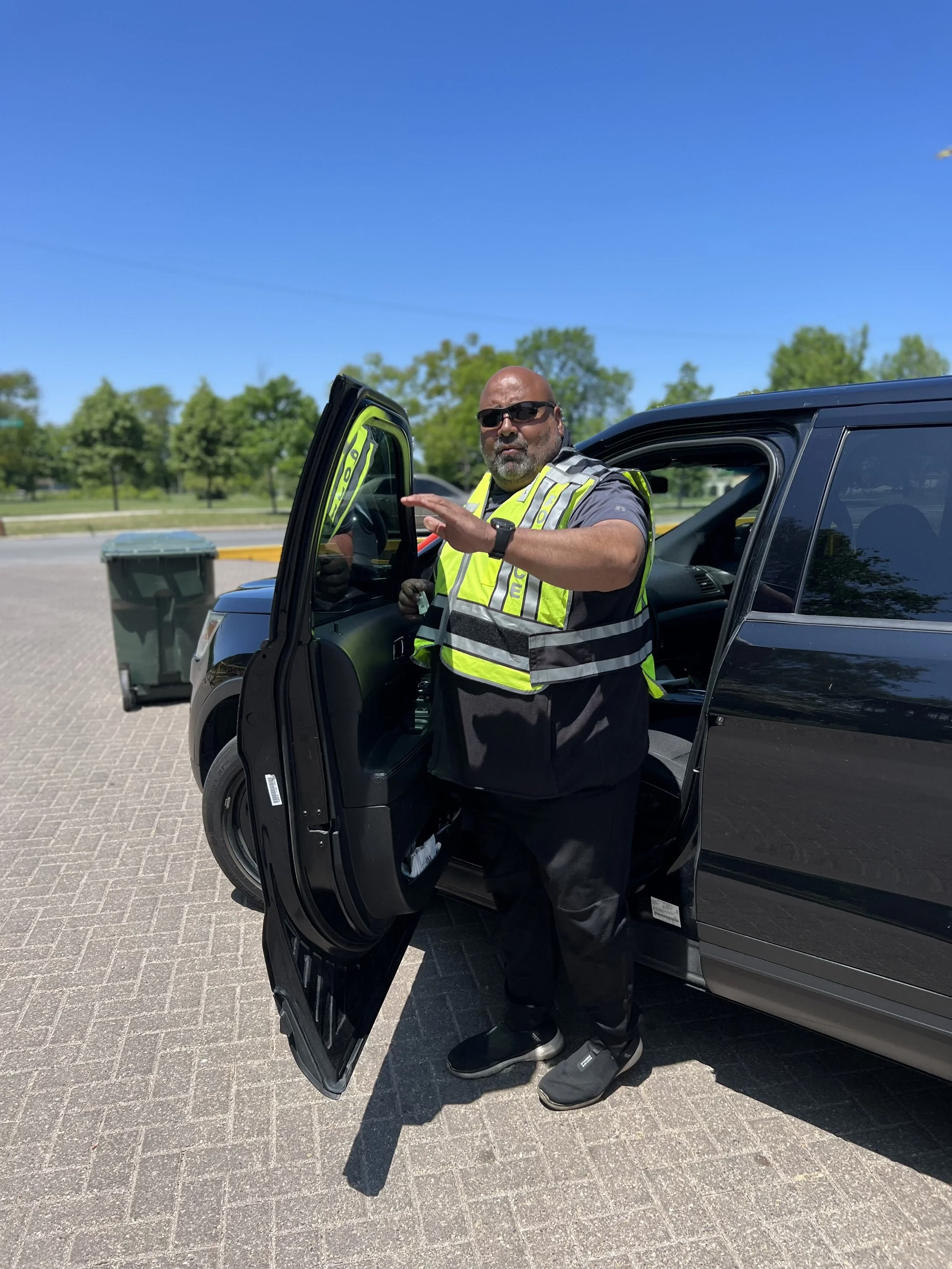 A security guard with sunglasses, a reflective safety vest, and gloves standing next to a tinted black pickup truck with its door open, in a parking lot with green trees in the background.