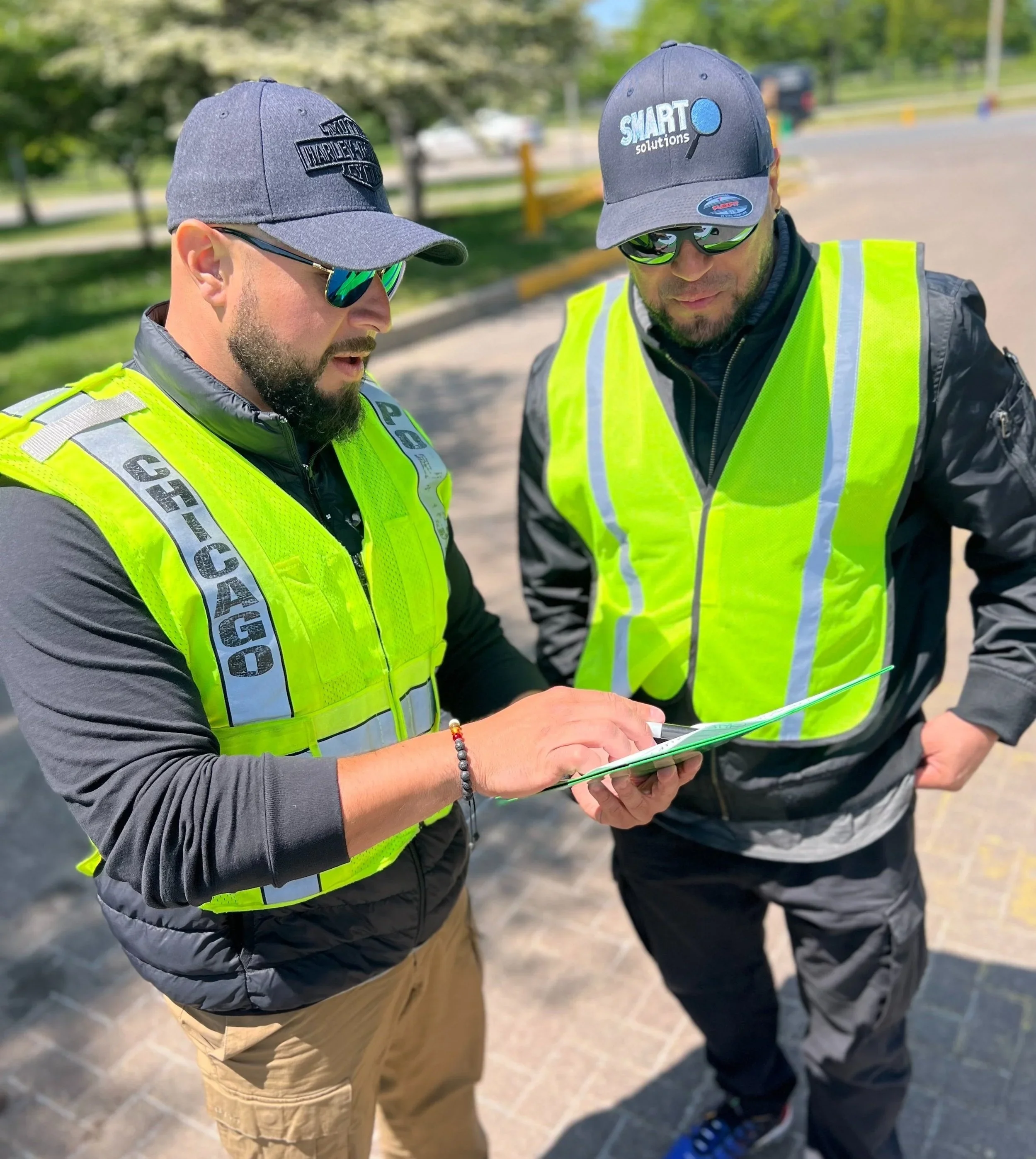 Two men wearing yellow safety vests and gray caps with logos are standing outside, looking at a paper or map. One man has sunglasses, a beard, and a bracelet; the other has a beard and glasses on his cap. The background shows trees and a parking lot.